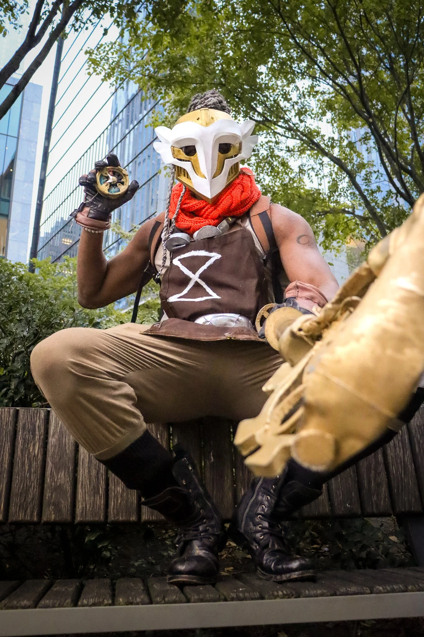 Person dressed as a warrior with a white and gold bird-like mask, red scarf, and brown clothing, sitting on a wooden bench outdoors in an urban area with skyscrapers and trees.