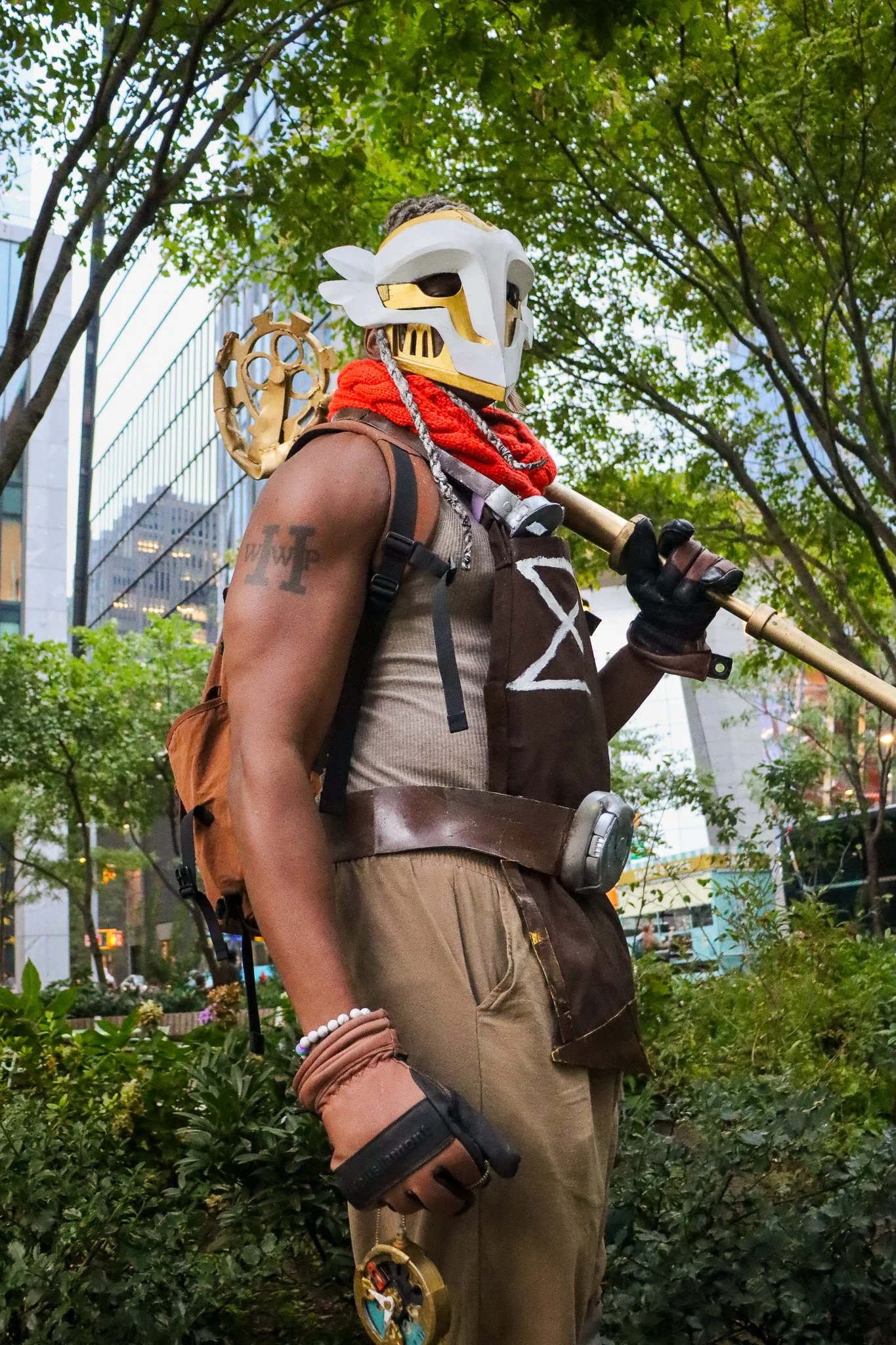A person wearing a bird skull mask, red scarf, and tactical gear, holding a staff in an urban park surrounded by trees and tall buildings.