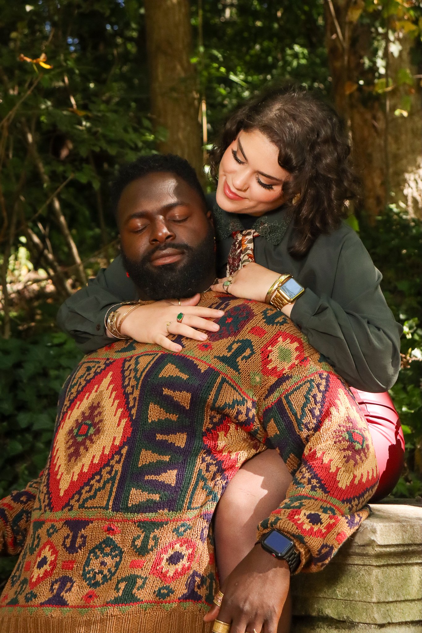 A woman with curly hair and light skin leans over and embraces a man with dark skin and a full beard sitting on a wooden bench in a forest. The woman is smiling and looking at the man, who has his eyes closed. The woman is wearing a dark green jacket