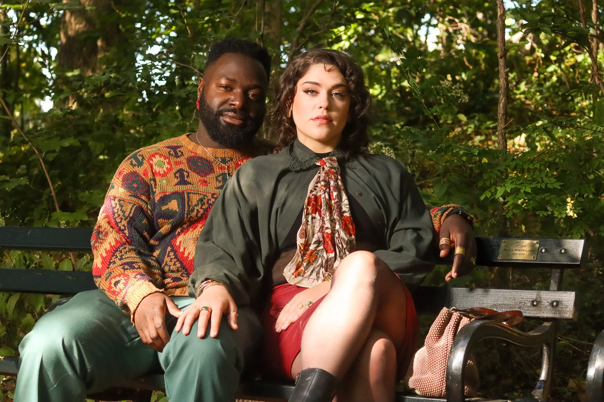 A man and woman sitting on a park bench surrounded by green foliage. The man and woman are celebrating their relationship with a couples photoshoot. They appear relaxed and contemplative, with the woman holding a small plaque or sign on the bench.