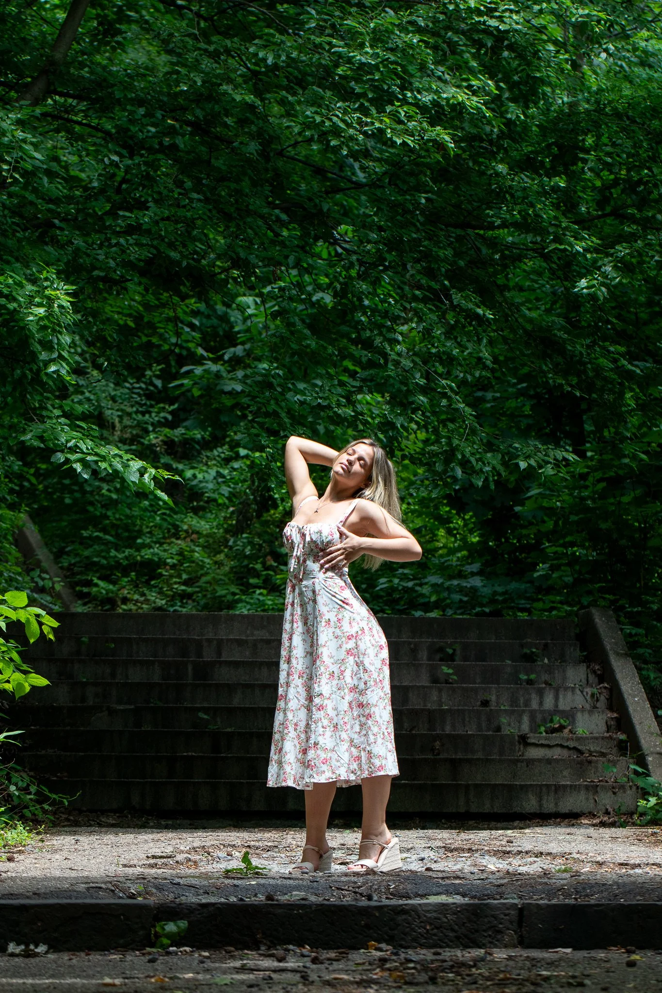 Woman in a floral dress standing on stone steps in a lush green forest, posing with one hand on her hip and the other behind her head.