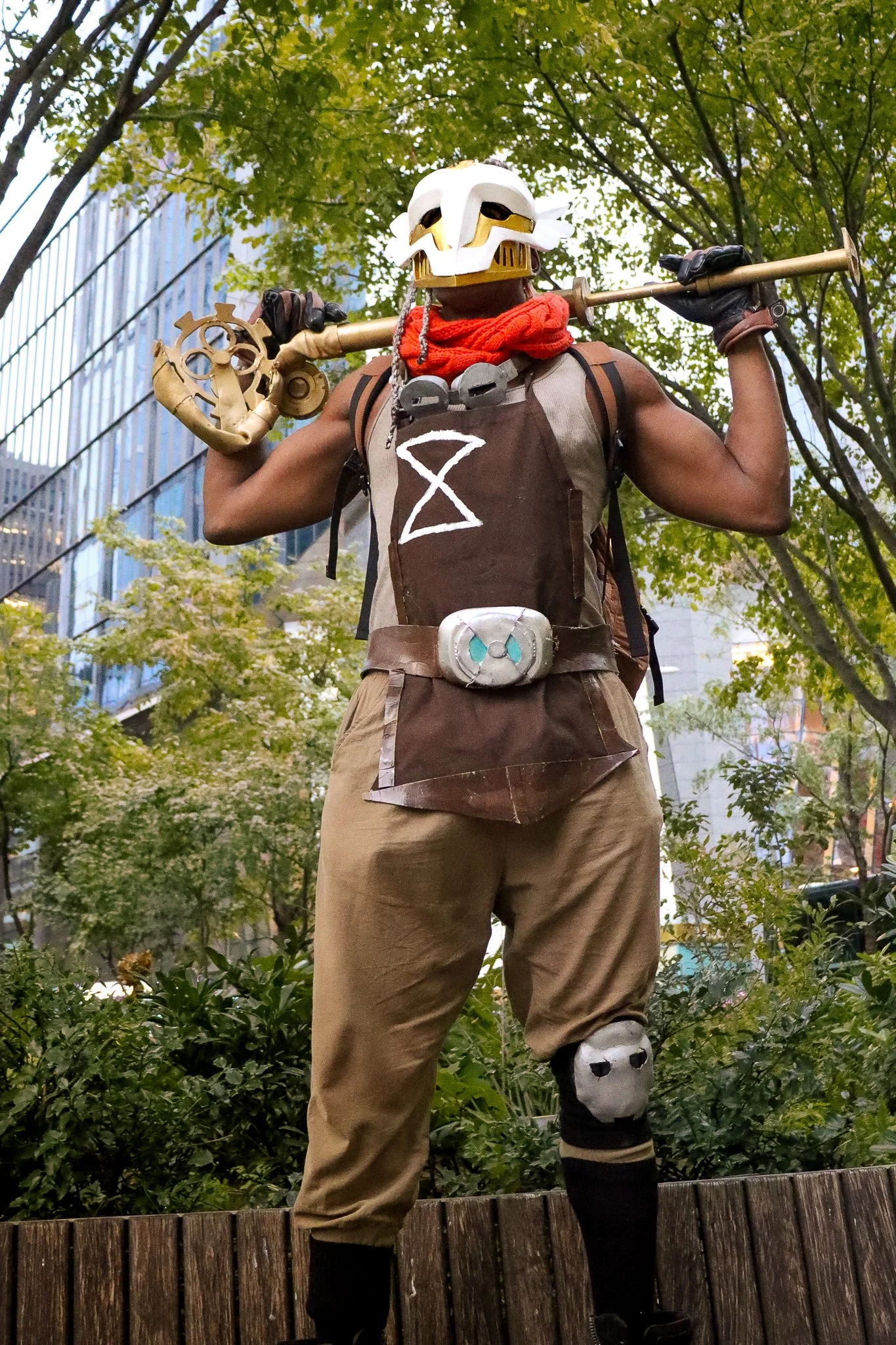 Costumed person standing outdoors with a tree and city buildings in the background, wearing a white skull mask, brown outfit, and holding a golden staff.