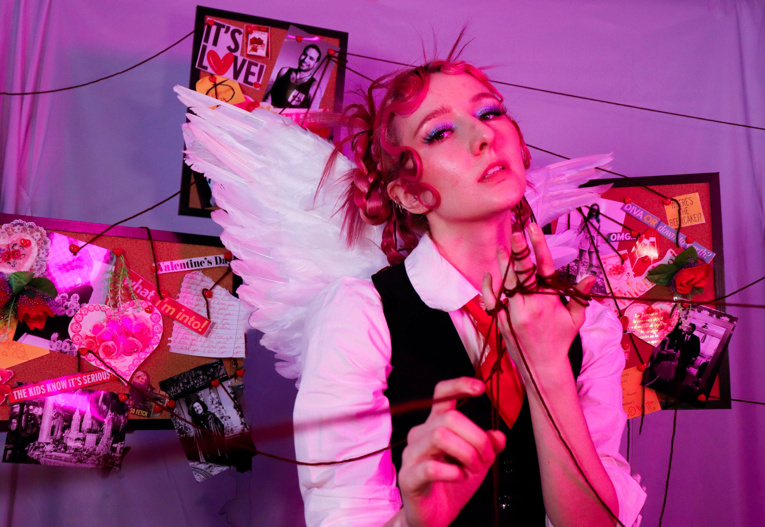 A person dressed as a cupid with pink curly hair, white wings, and a white shirt, holding a bow with a heart-shaped arrow in a decorated Valentines Day-themed room.