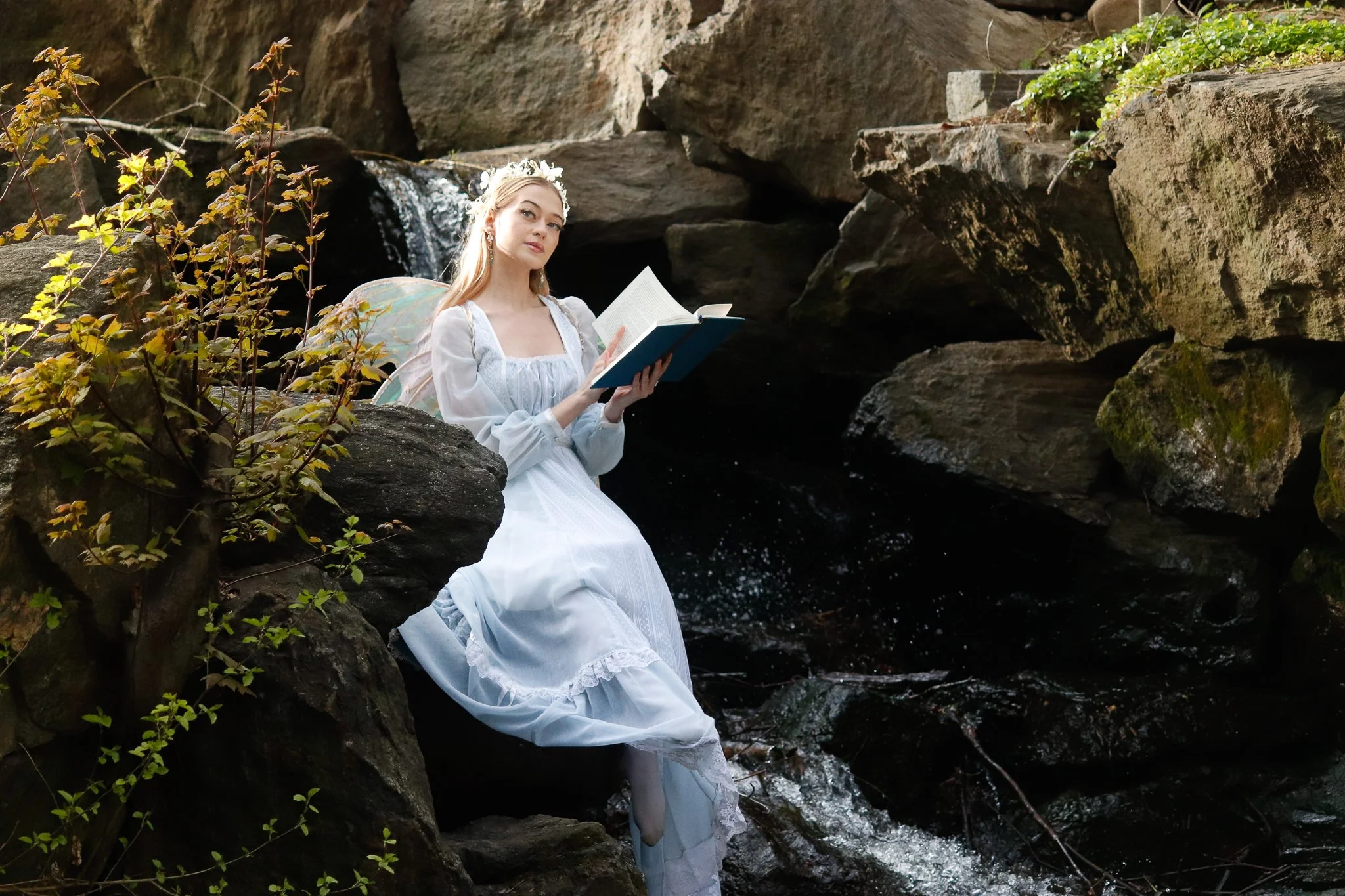 A young woman dressed as a fairy with wings and a white dress sitting on rocks by a small waterfall, holding a book, surrounded by green plants and rocks.