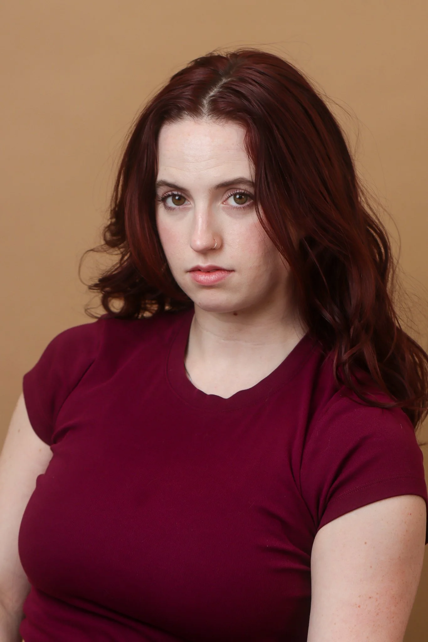 A young woman with red, wavy hair, wearing a maroon shirt, looking directly at the camera against a plain beige background.