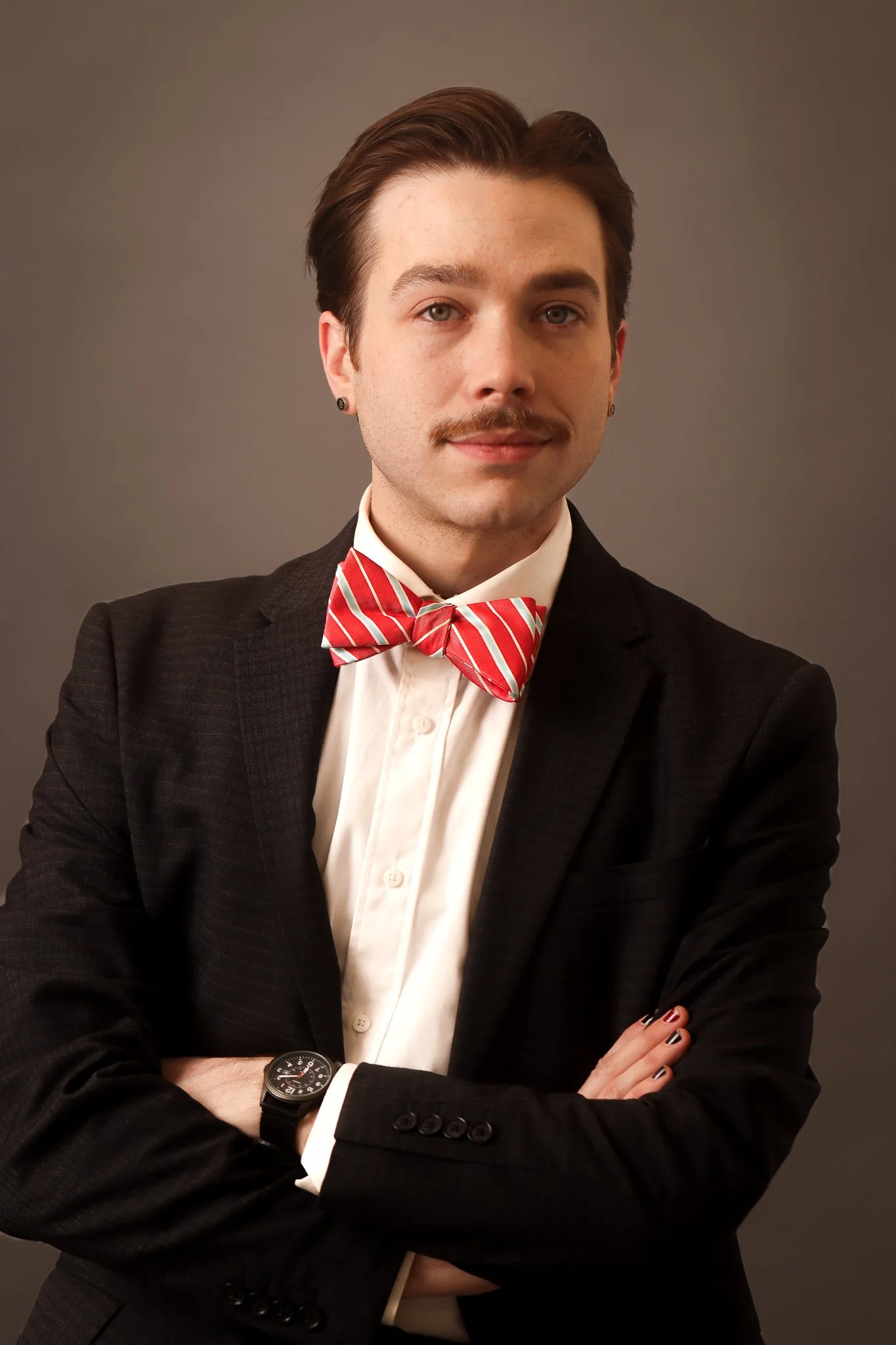 A young man with dark hair, mustache, and light-colored eyes wearing a black suit, white dress shirt, red and white striped bow tie, and a black wristwatch, with arms crossed against a plain gray background.