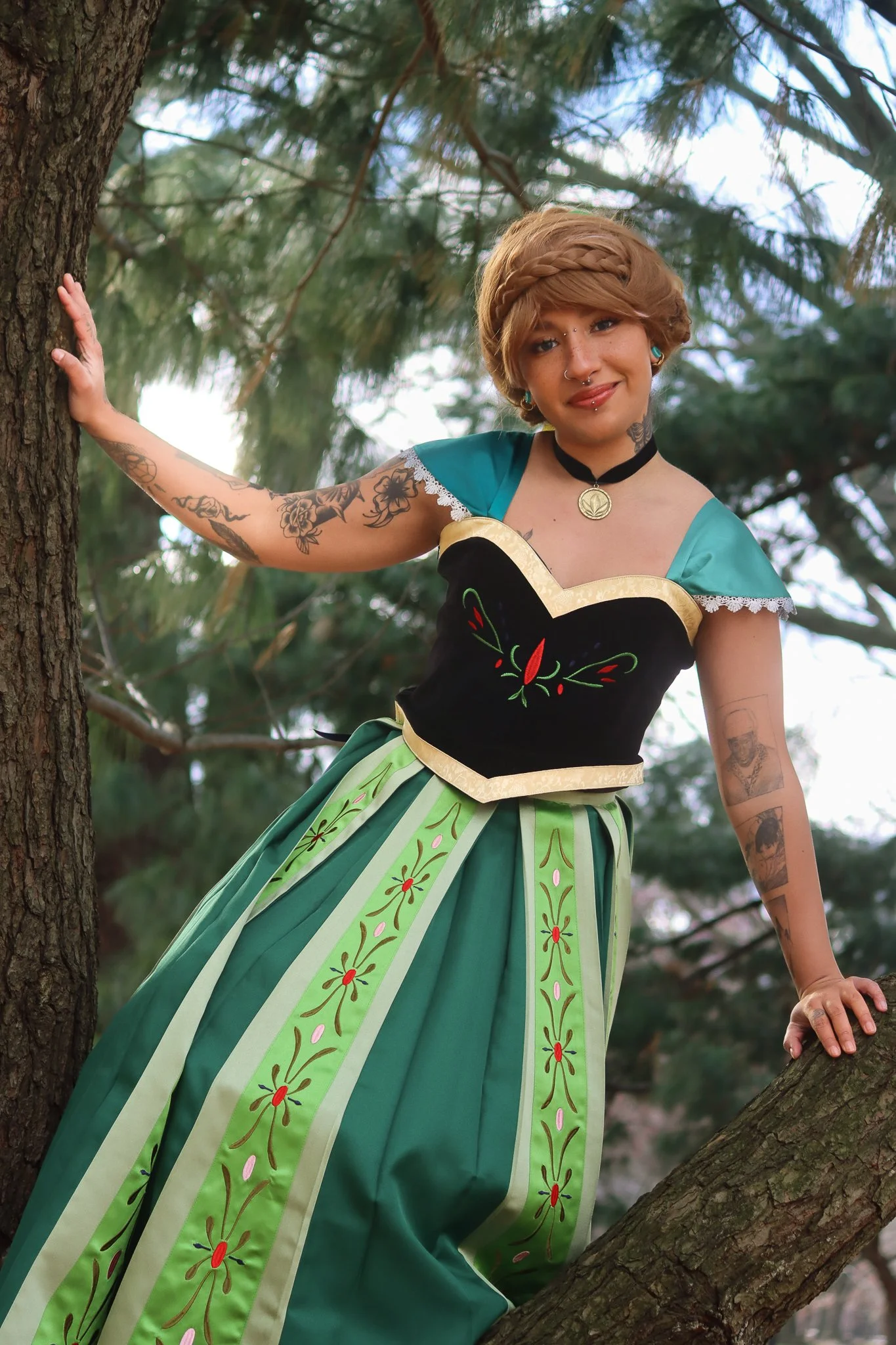 A woman with short, wavy, light brown hair, tattoos on her arms, and multiple facial piercings, standing on a tree and smiling. She is wearing a traditional dress with green and black colors and floral embroidery, and a black choker with a pendant. The background features pine tree branches.