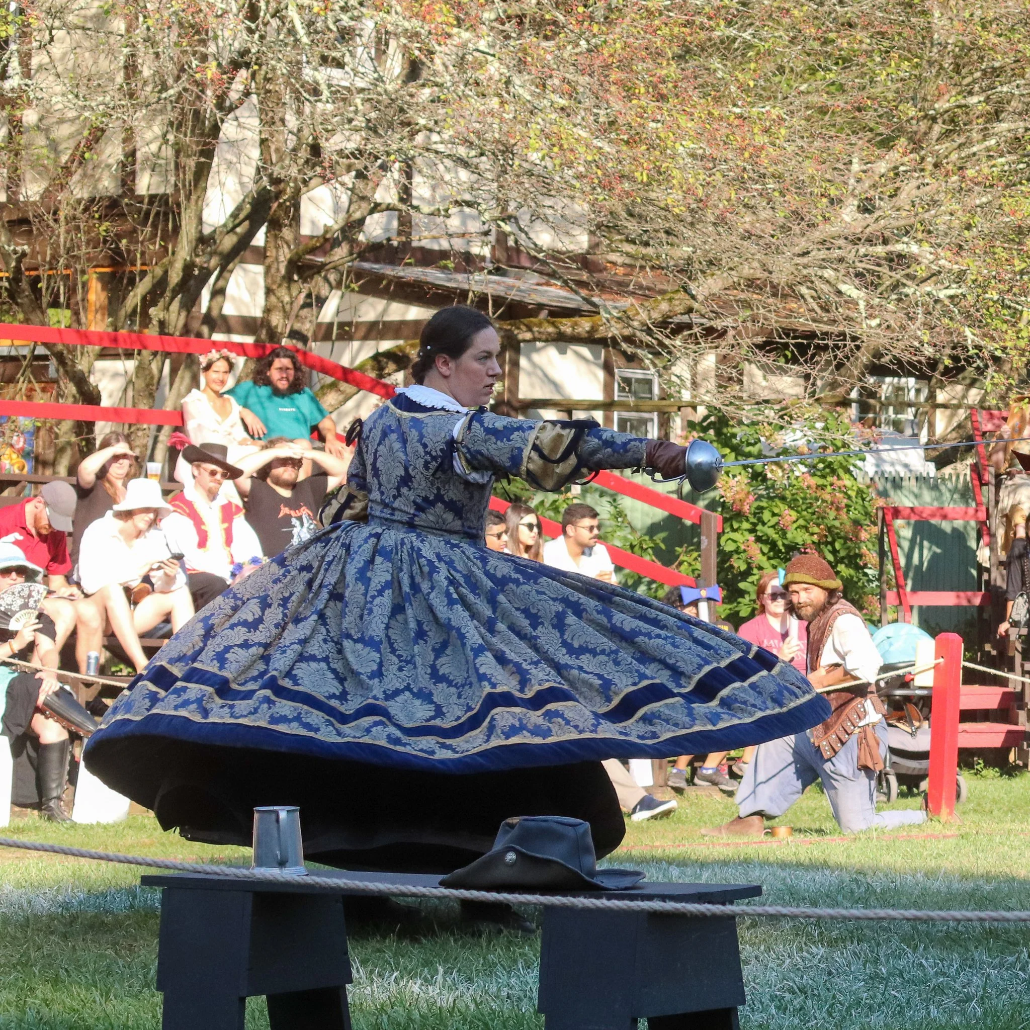 A woman dressed in historical costume riding a horse in front of a crowd at an outdoor event surrounded by trees and spectators.