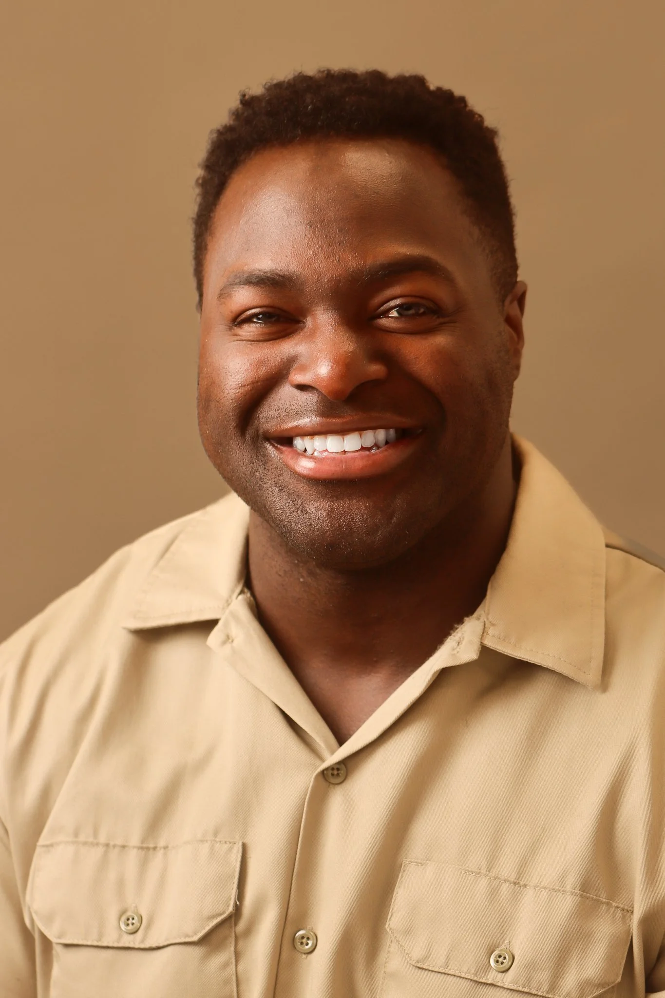 Close-up portrait of a smiling Black man with short, curly hair wearing a beige collared shirt against a plain background.