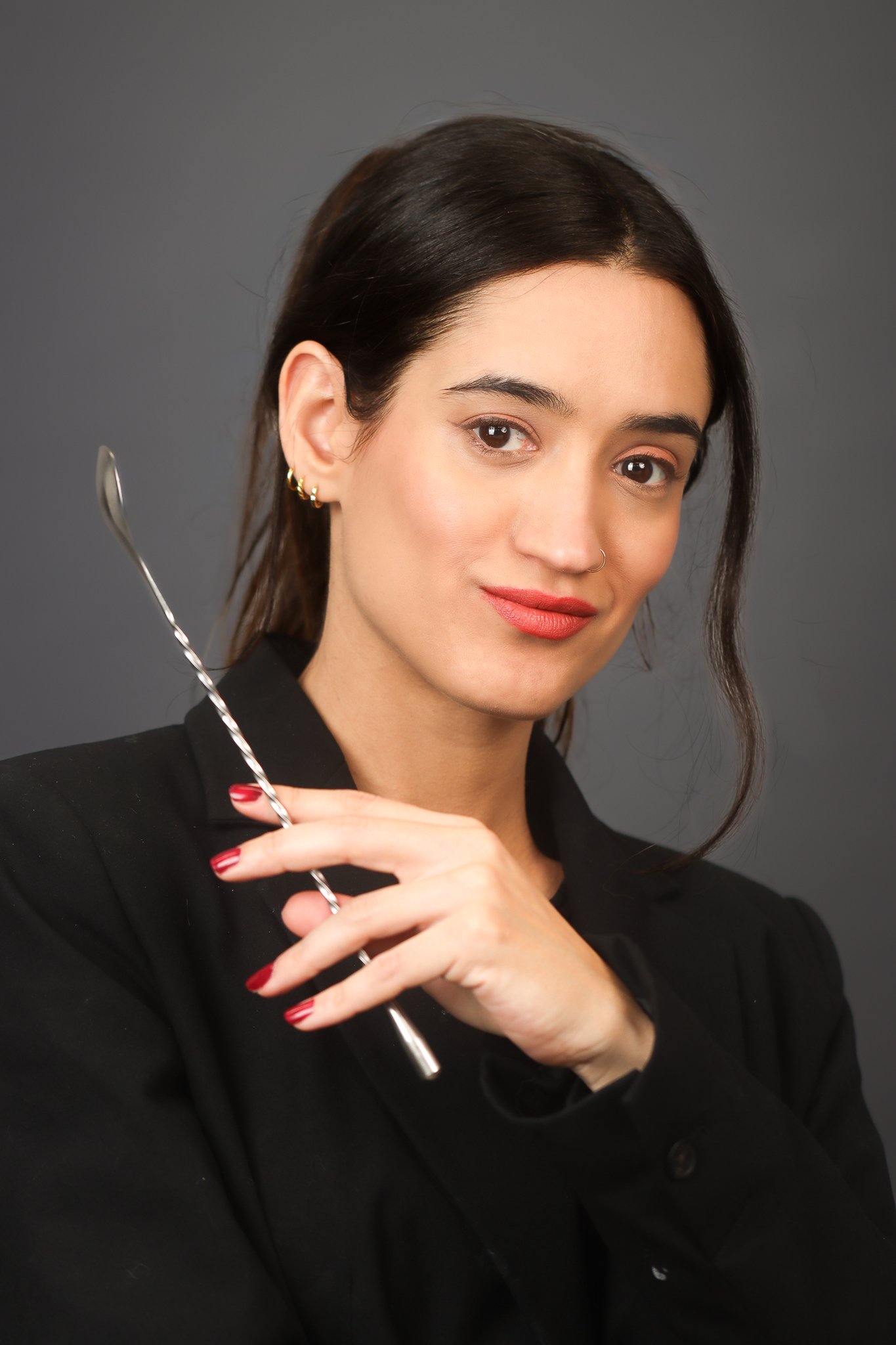 A woman with dark hair, wearing gold earrings and a nose ring, holds a metal stirrer. She is wearing a black outfit and has red nail polish, set against a plain gray background.