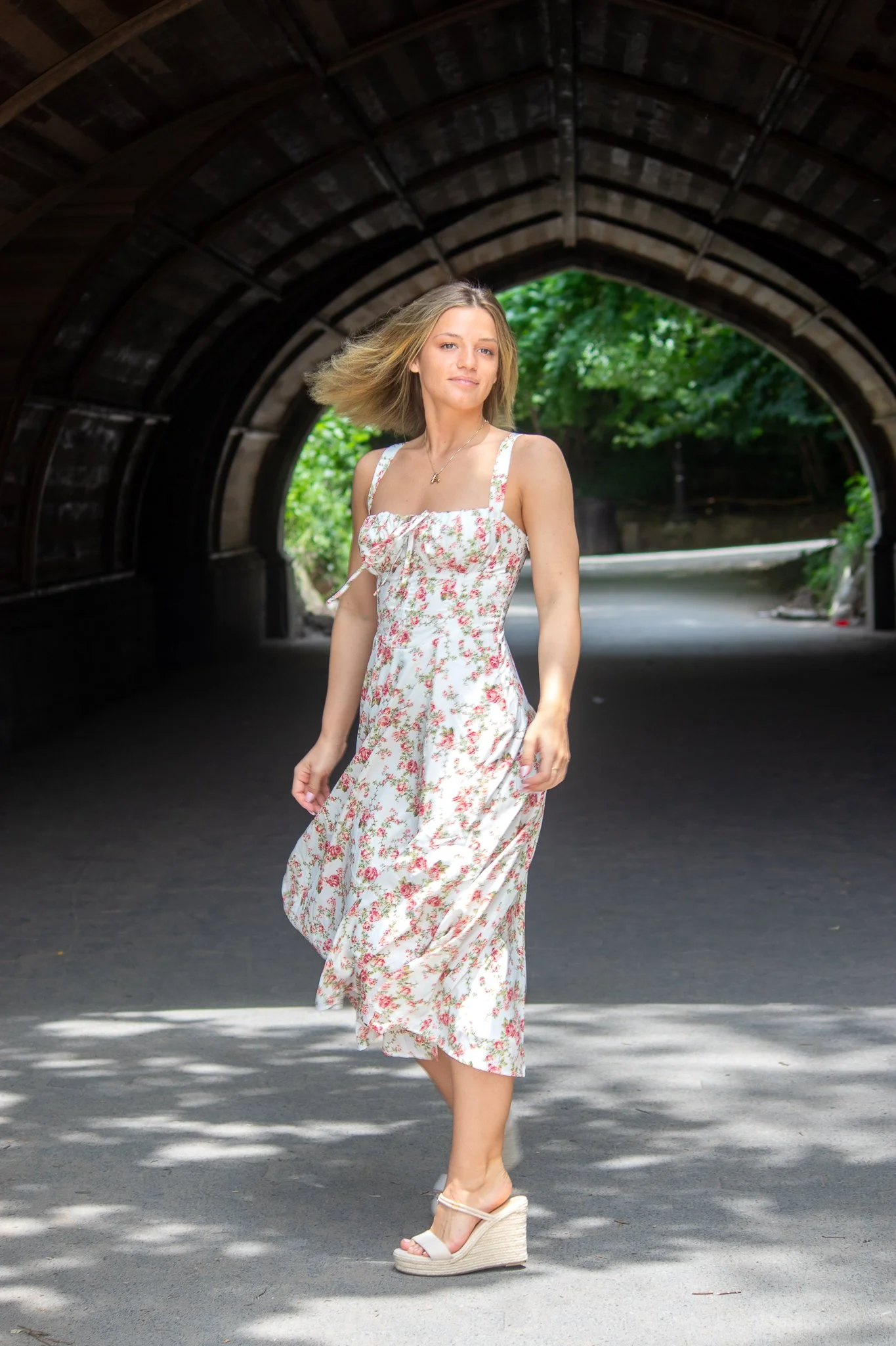 A woman with blonde hair wearing a white floral dress and wedge sandals walking under a tunnel with a green, leafy background.