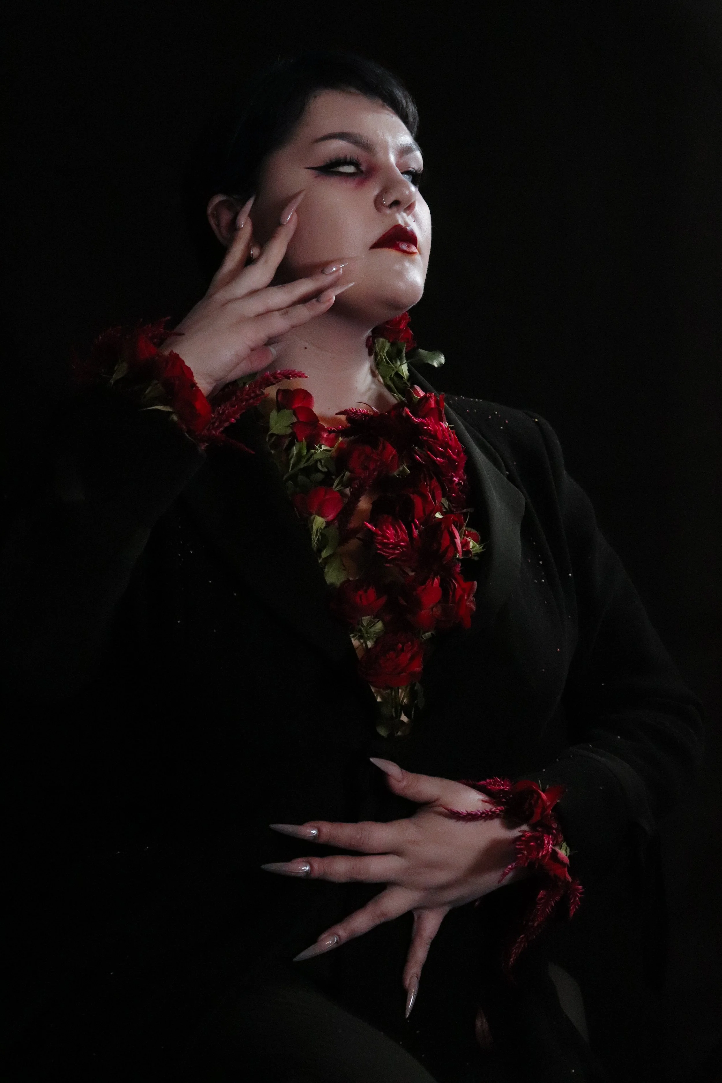 A woman with dark hair and makeup, wearing red flowers and black clothing, poses against a dark background.