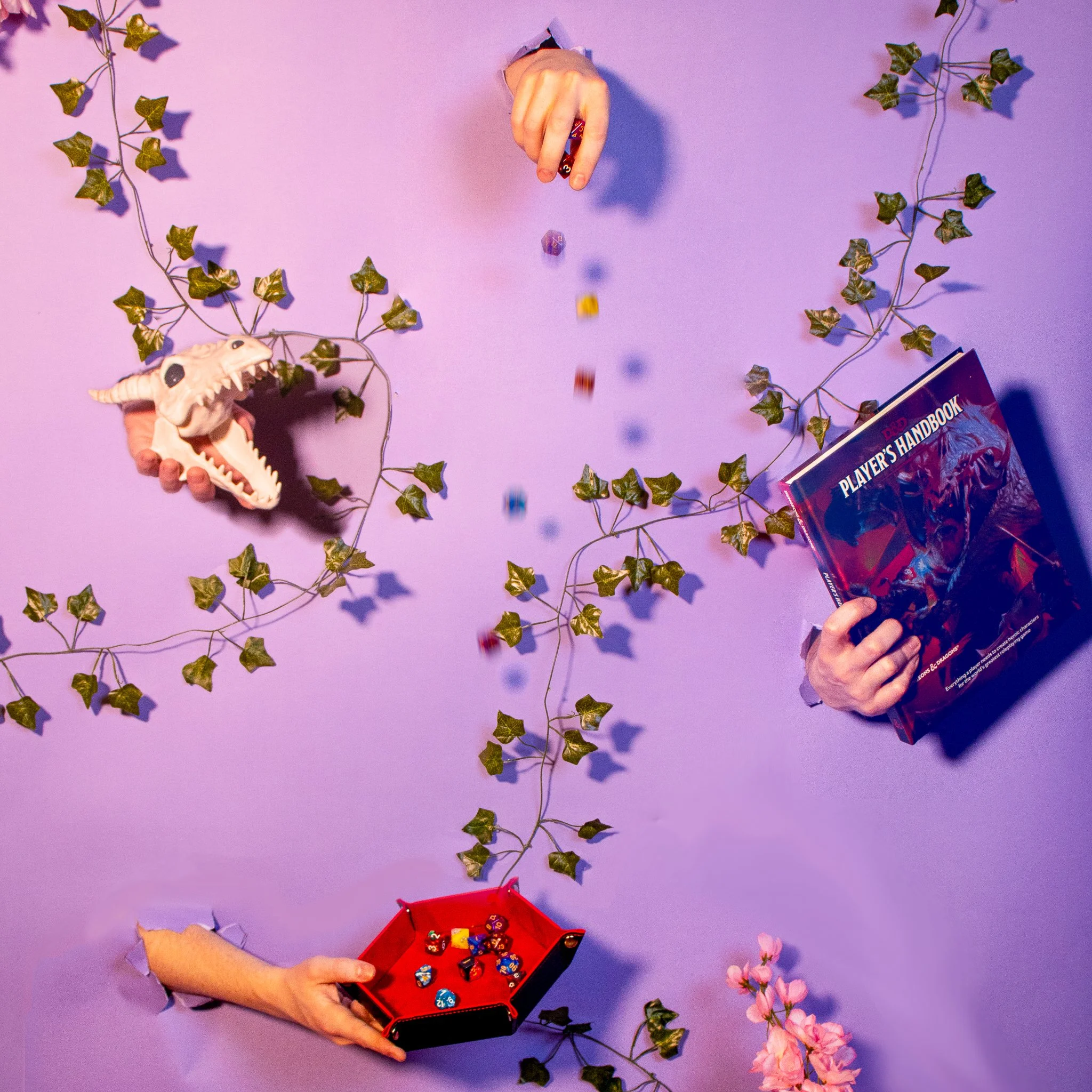 People playing a tabletop role-playing game with a red box containing colorful dice on a purple background decorated with artificial green ivy and pink flowers, with torn paper edges on their sleeves.