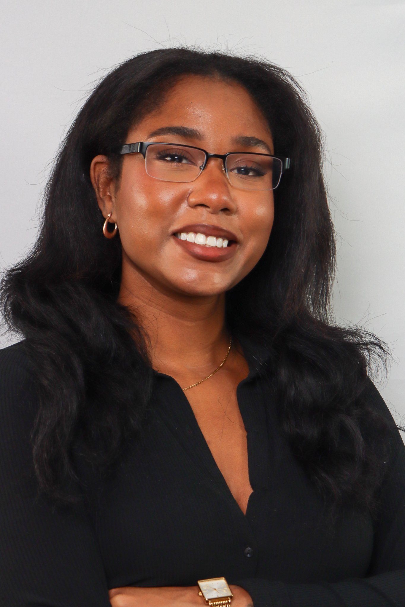 A woman with dark hair, glasses, and hoop earrings smiling at the camera, wearing a black top and a gold watch.