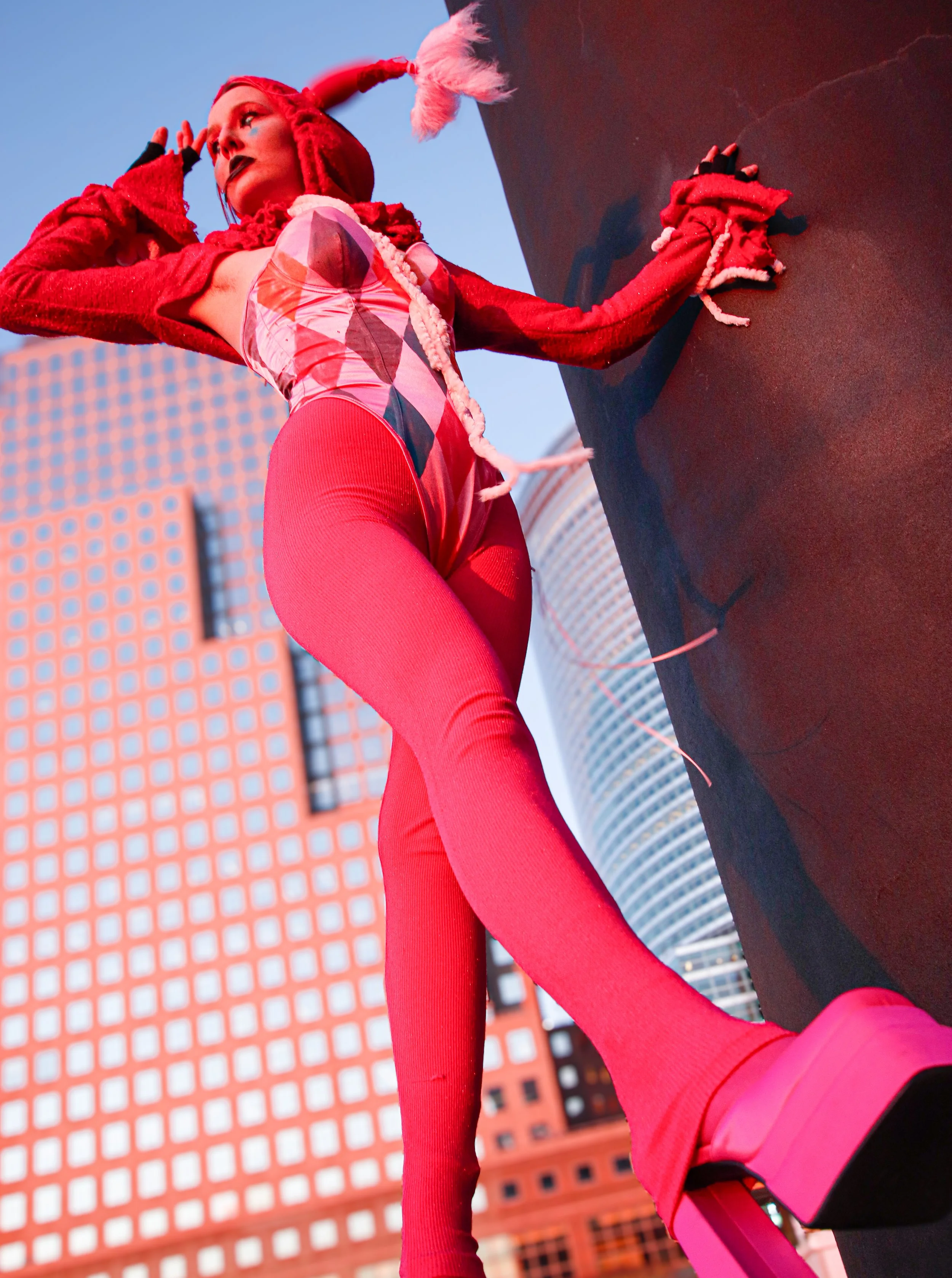 Person dressed in vibrant red and pink clothing, including a hoodie with a feathered tip and matching leggings, standing against a cityscape background with tall buildings and a clear blue sky.