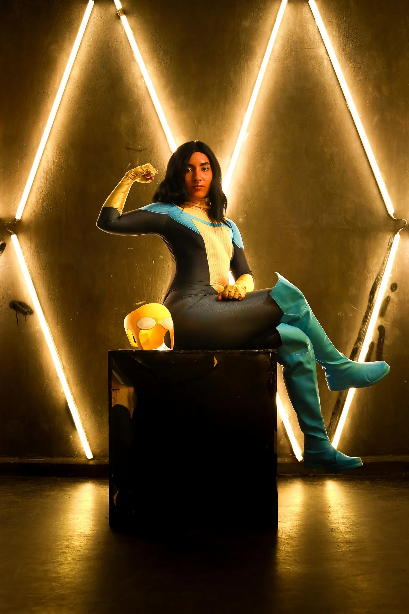 Woman dressed as a superhero sitting on a black box with her arm flexed, wearing a colorful costume with blue, yellow, and black, with a yellow helmet with ear holes placed on the box, in front of a metallic wall with neon lights forming geometric pa