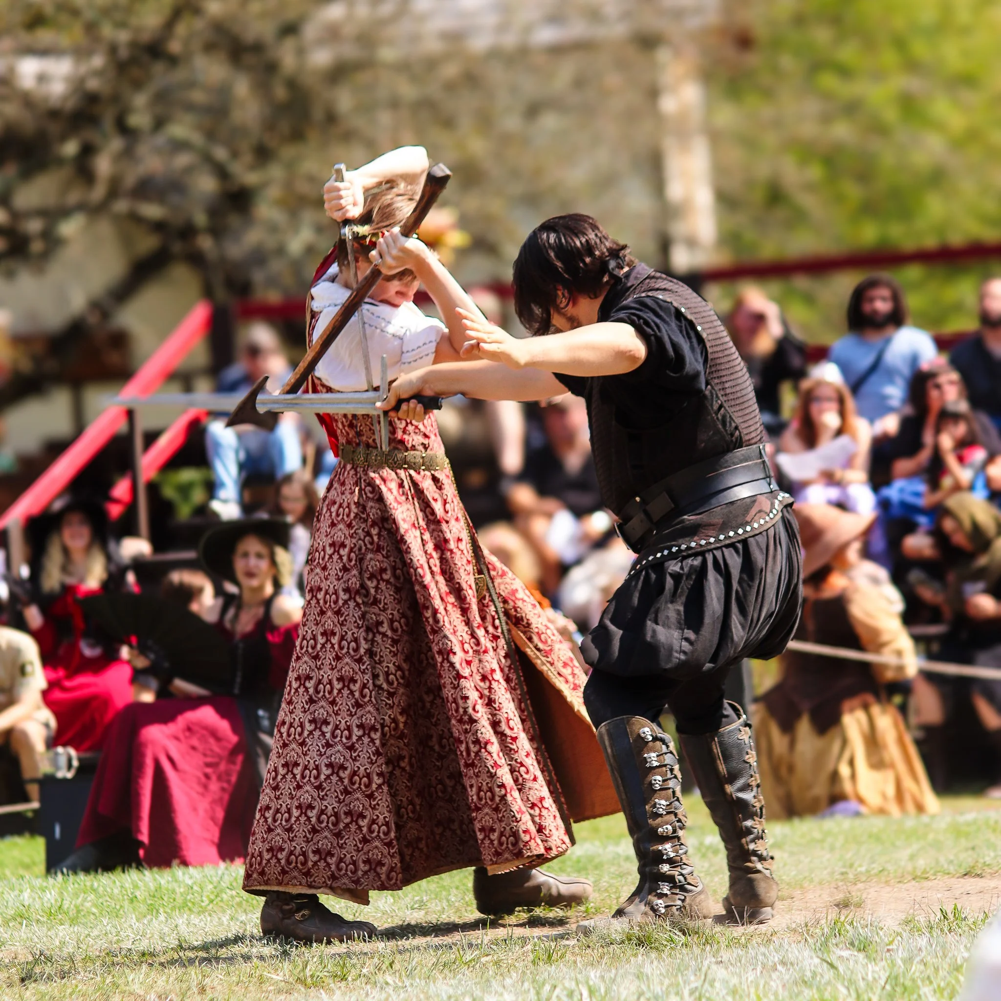 Two people, dressed in medieval or fantasy costumes, perform a sword fight on a grassy area in front of an audience. The woman wears a long, patterned skirt and a blouse, while the man wears black leather armor and tall black boots.