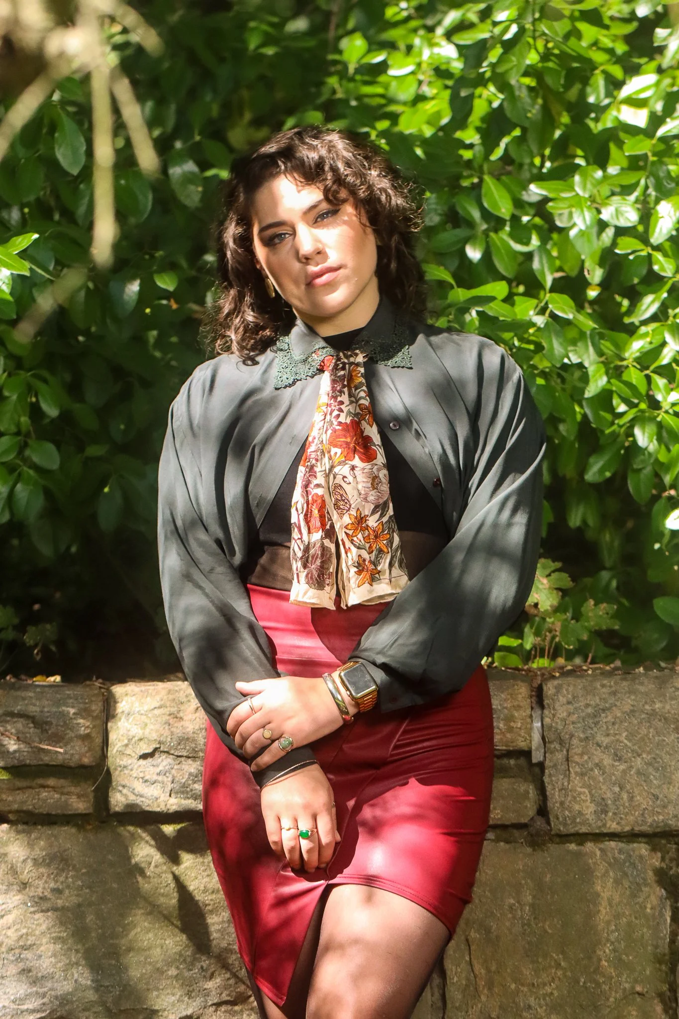 A woman with curly dark hair standing outdoors in front of green bushes, wearing a black blouse with lace collar, a patterned scarf, a red skirt, and accessorized with rings, bracelets, and a smartwatch.