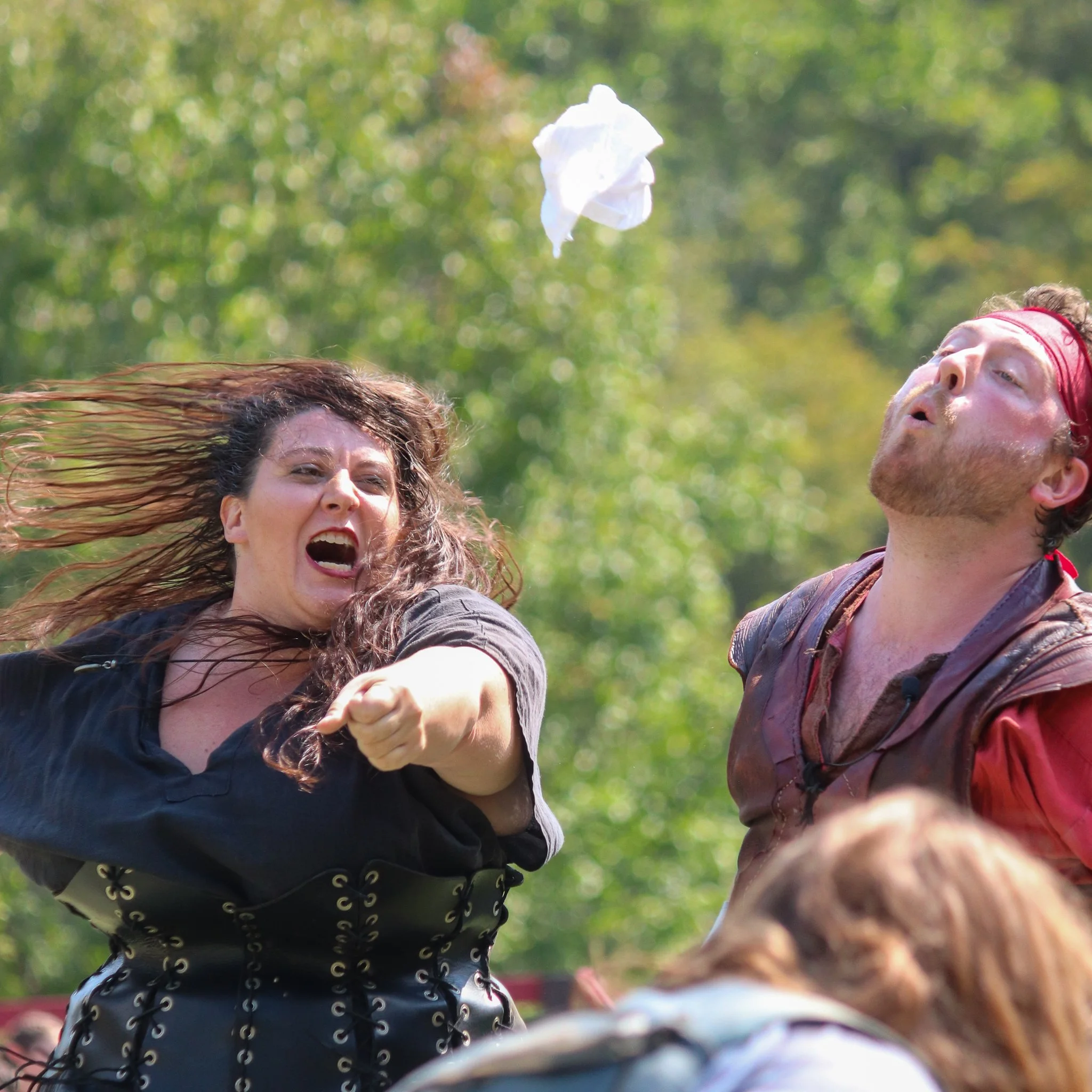 Group of people celebrating outdoors, some jumping with excitement, one woman smiling with her eyes closed and mouth open, a man wearing a red headband, blurred background of green trees.