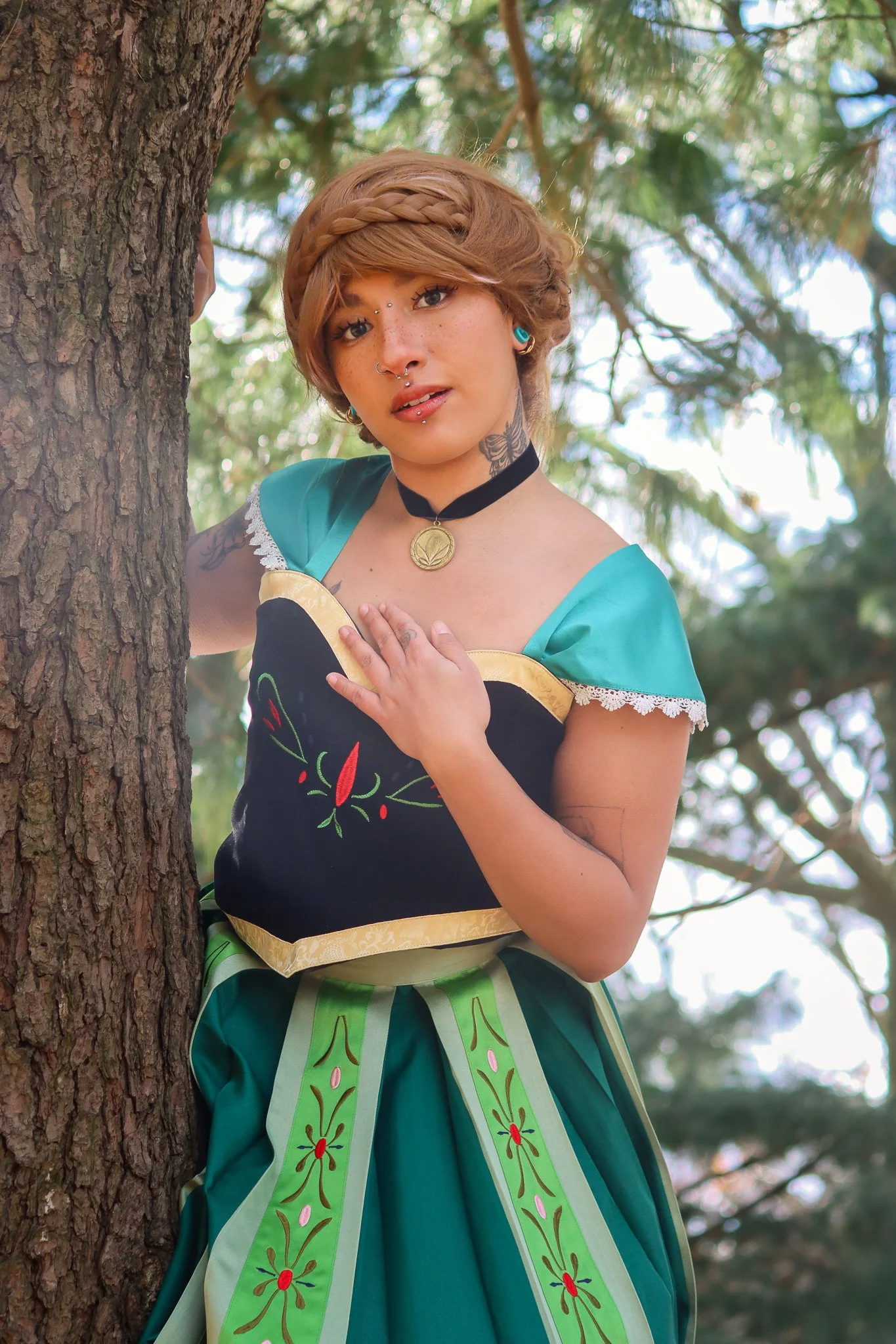 Young woman with short, wavy, blonde hair styled with braids, wearing a dress with green, black, and yellow colors, a black choker with a pendant, and standing outdoors near a tree with branches and leaves in the background.