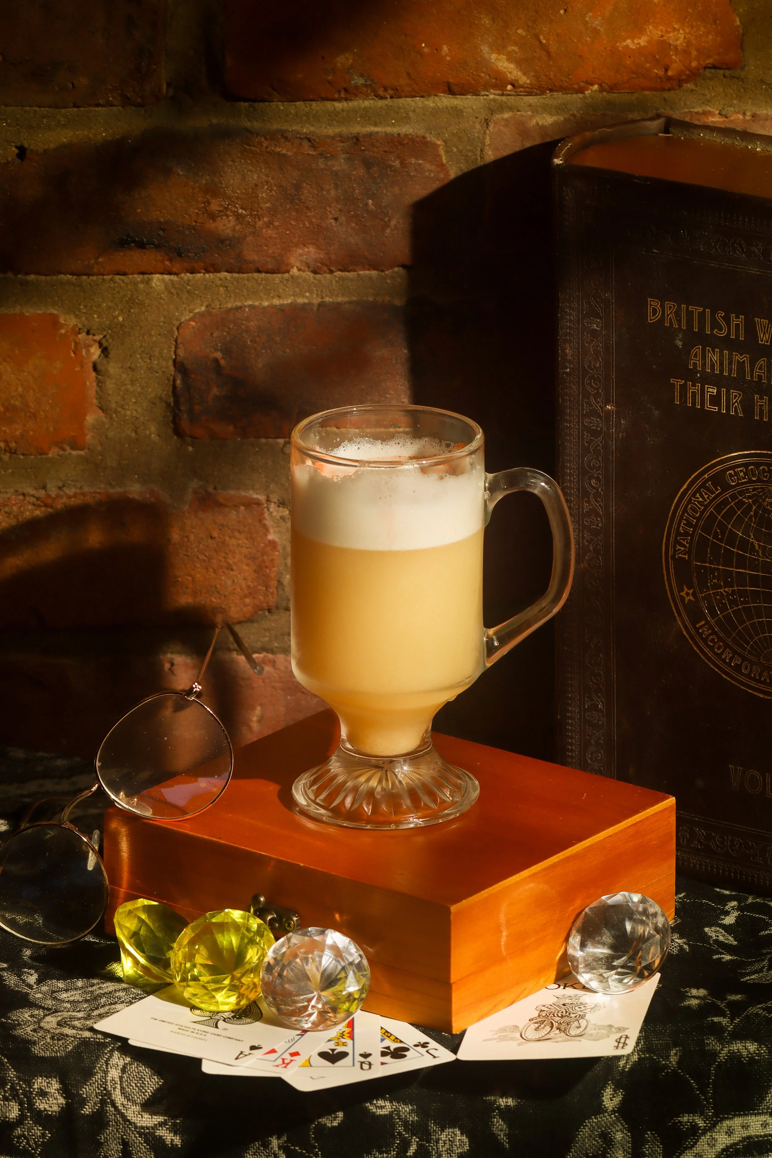 A glass of beer, a deck of playing cards, sunglasses, and decorative gemstones on a table with a brick wall background.