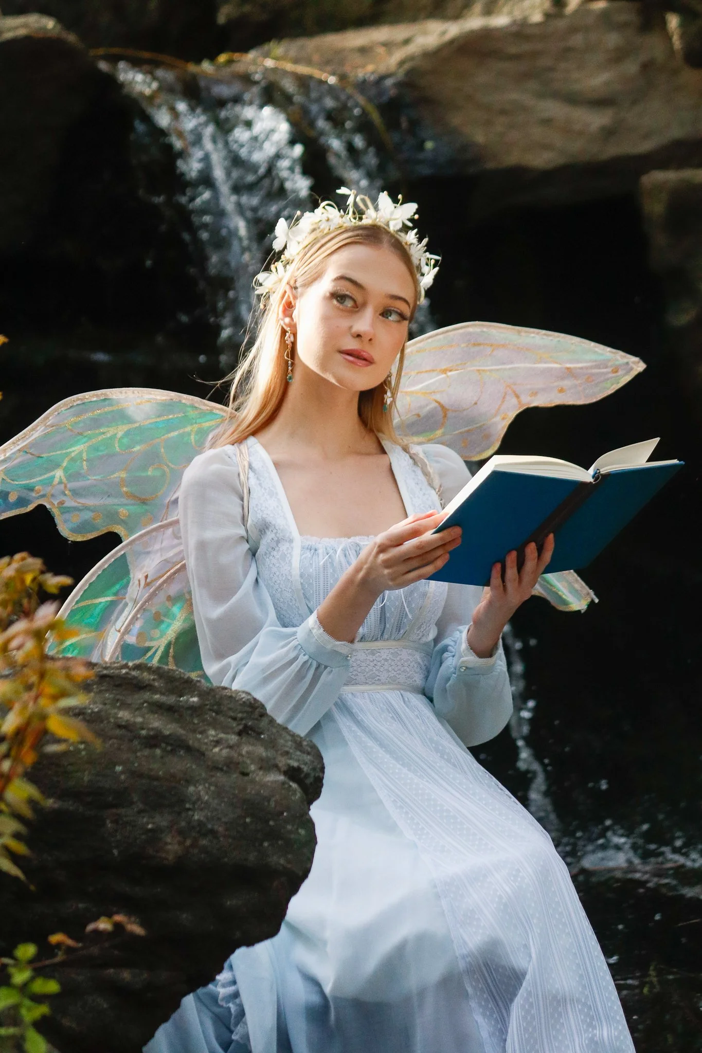 A woman dressed as a fairy with large, iridescent wings, wearing a flower crown and holding an open book, sitting outdoors near a waterfall.