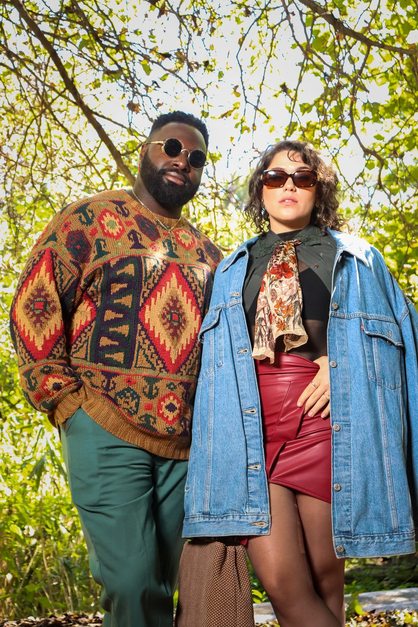 A man and woman standing outdoors among green trees, both wearing sunglasses. The man has a beard and is wearing a colorful patterned sweater with green pants. The woman has curly hair and is wearing an oversized denim jacket, a black top, red leathe