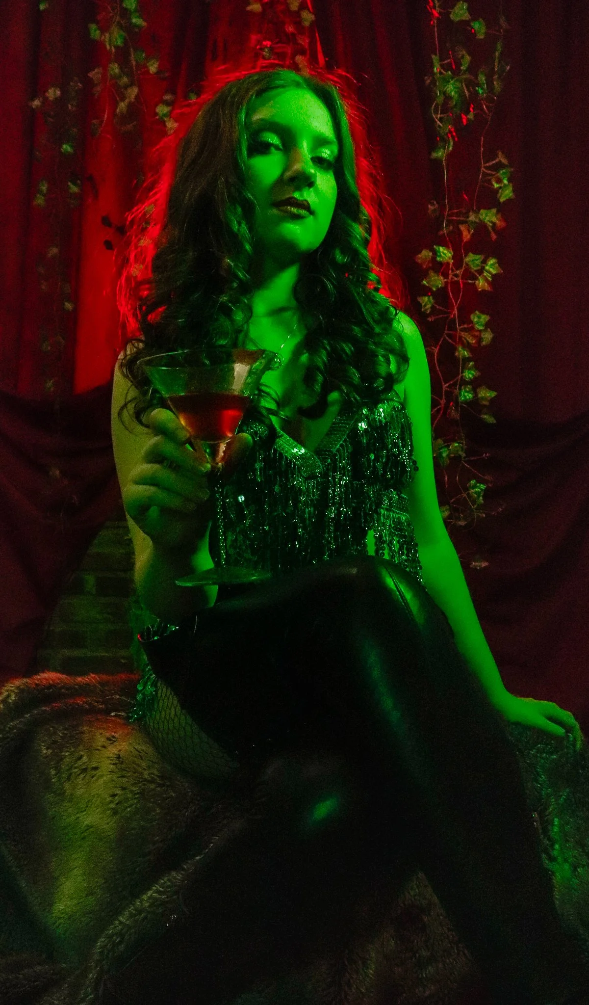 A woman with dark curly hair, dark lipstick, and eye makeup, holding a martini glass filled with a dark red drink, sitting in a dimly lit room with dark red curtains and vine decorations, illuminated by green and red lighting.