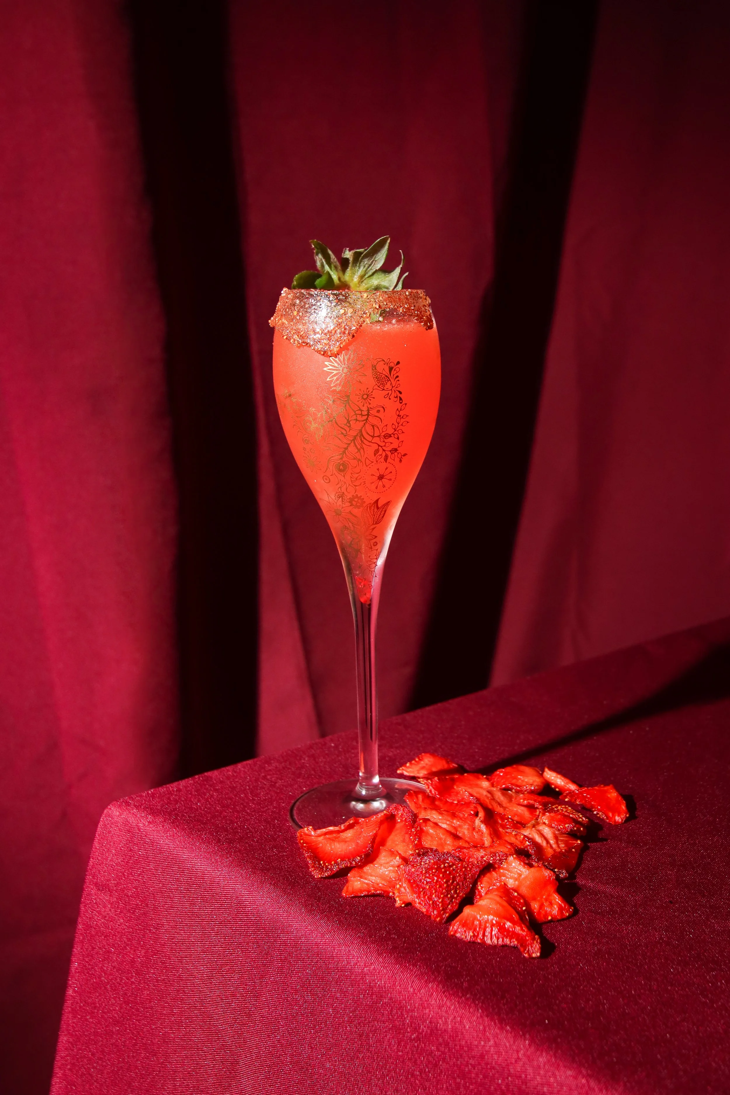 A tall glass with a strawberry cocktail garnished with a strawberry, glittery sugar rim, and decorative gold design, placed on a red tablecloth with sliced strawberries underneath.