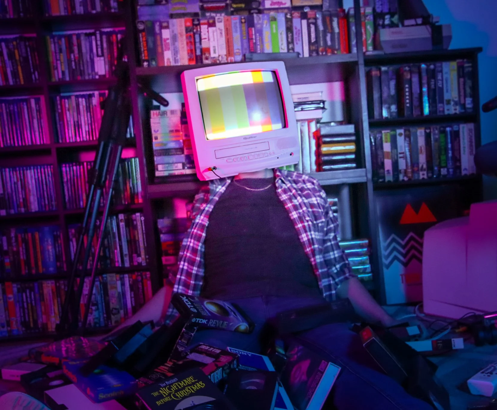 A person sitting in a room with shelves filled with books in the background. The person has a vintage CRT TV as a head displaying a test pattern. The person is wearing a plaid shirt and black t-shirt, surrounded by scattered boxes and electronics, wi