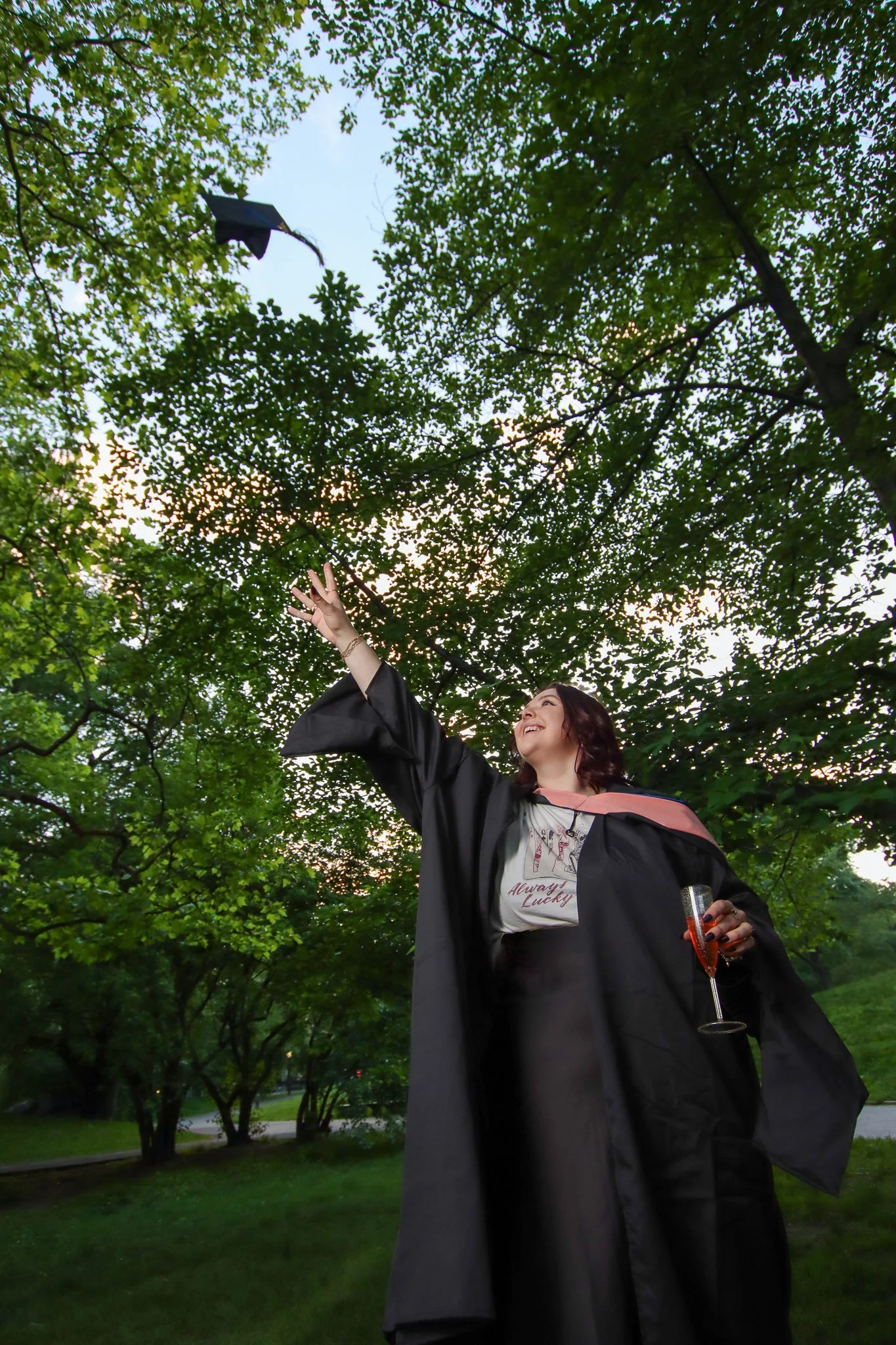 A woman in a graduation gown and cap celebrating outdoors, holding a glass of champagne with trees and a blue sky in the background.