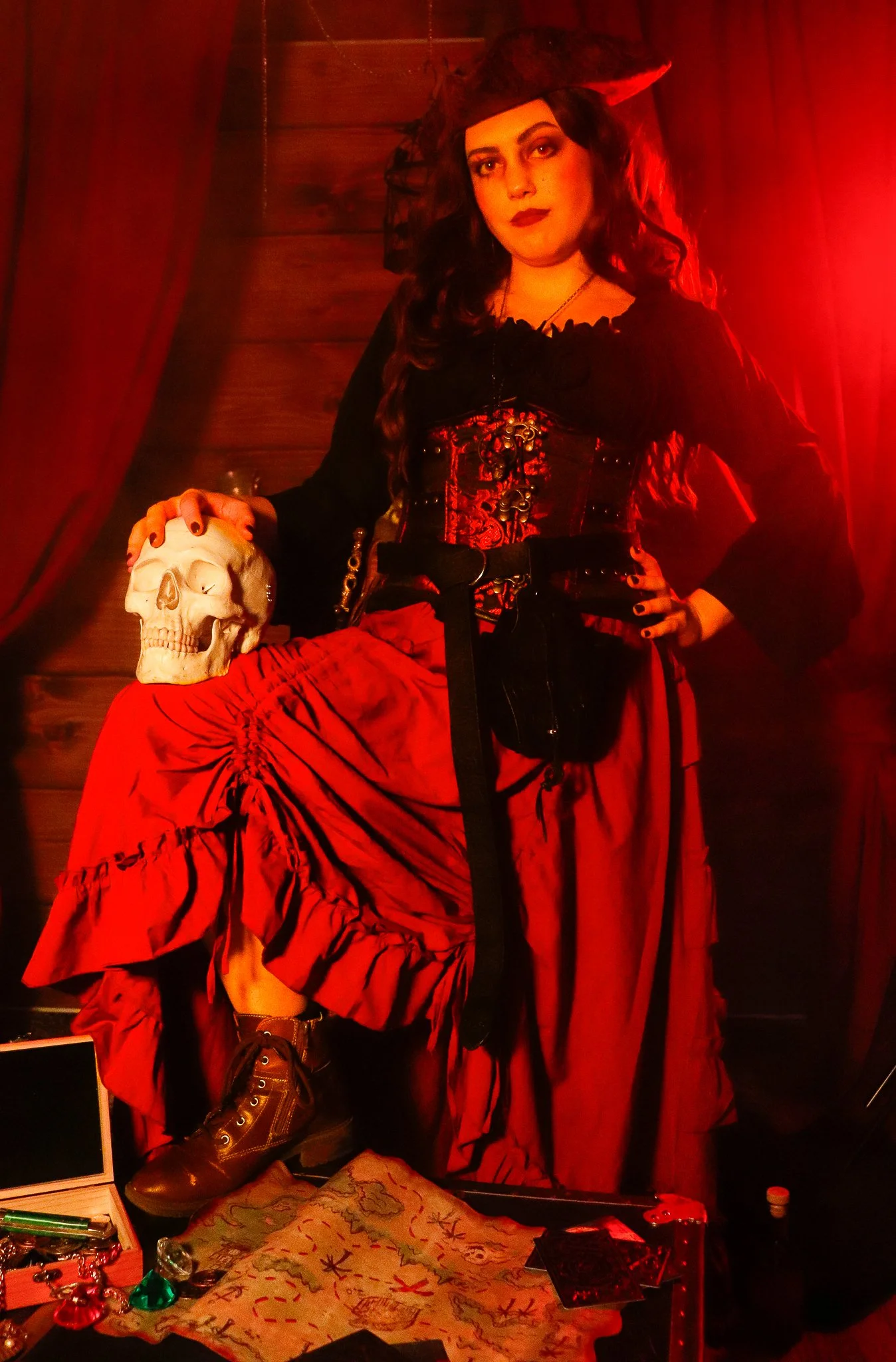 A woman dressed as a pirate with a red and black costume, pirate hat, and long dark hair, standing in front of a red curtain, holding a human skull.