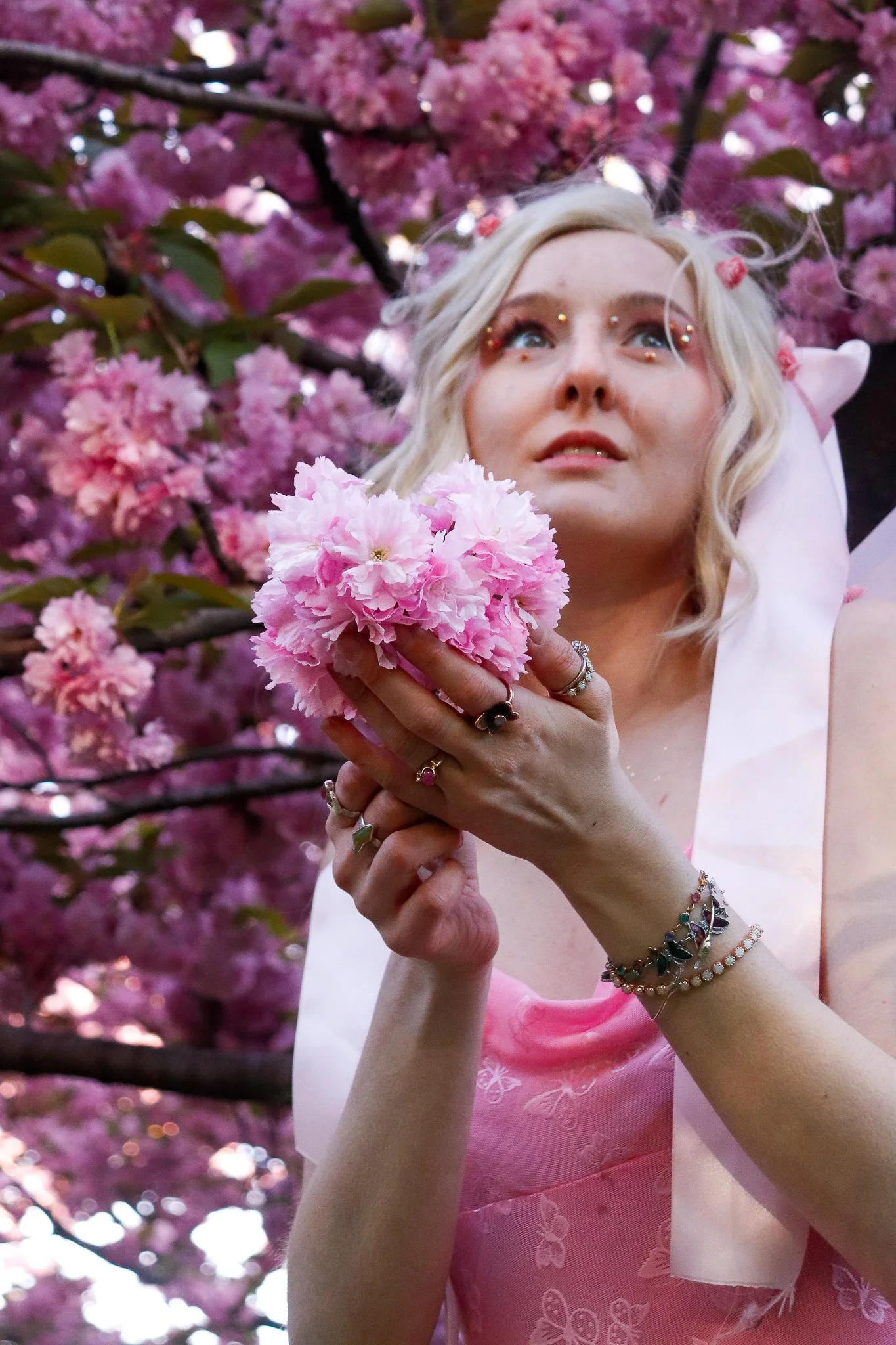 A woman with blonde hair and blue eyes holding a pink flower, wearing a pink dress and multiple jewelry bracelets, surrounded by pink blossoms on a tree.