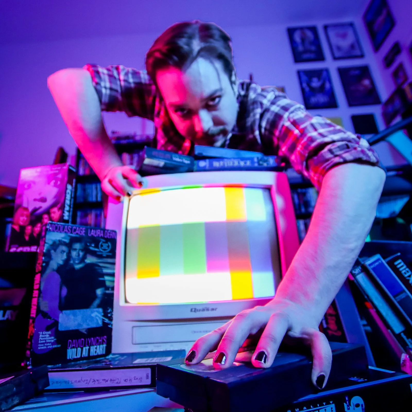 A person with dark hair and plaid shirt leaning over an old CRT television with a color test screen. Hands with dark nail polish are on the TV, surrounded by books, DVDs, and framed pictures on the wall.