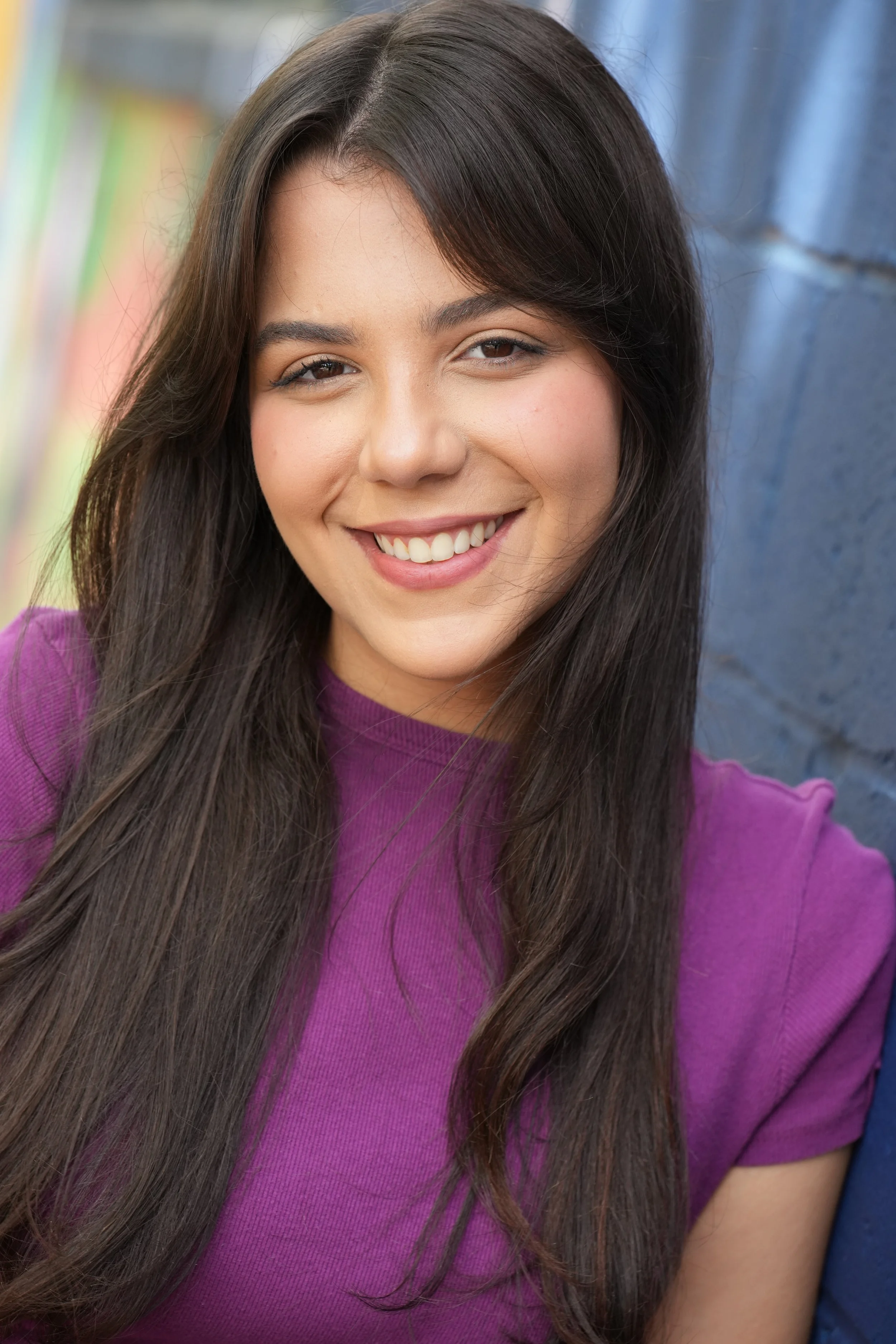Close-up of a young woman with long dark hair, smiling, wearing a purple top, with a blue wall and colorful books in the background.