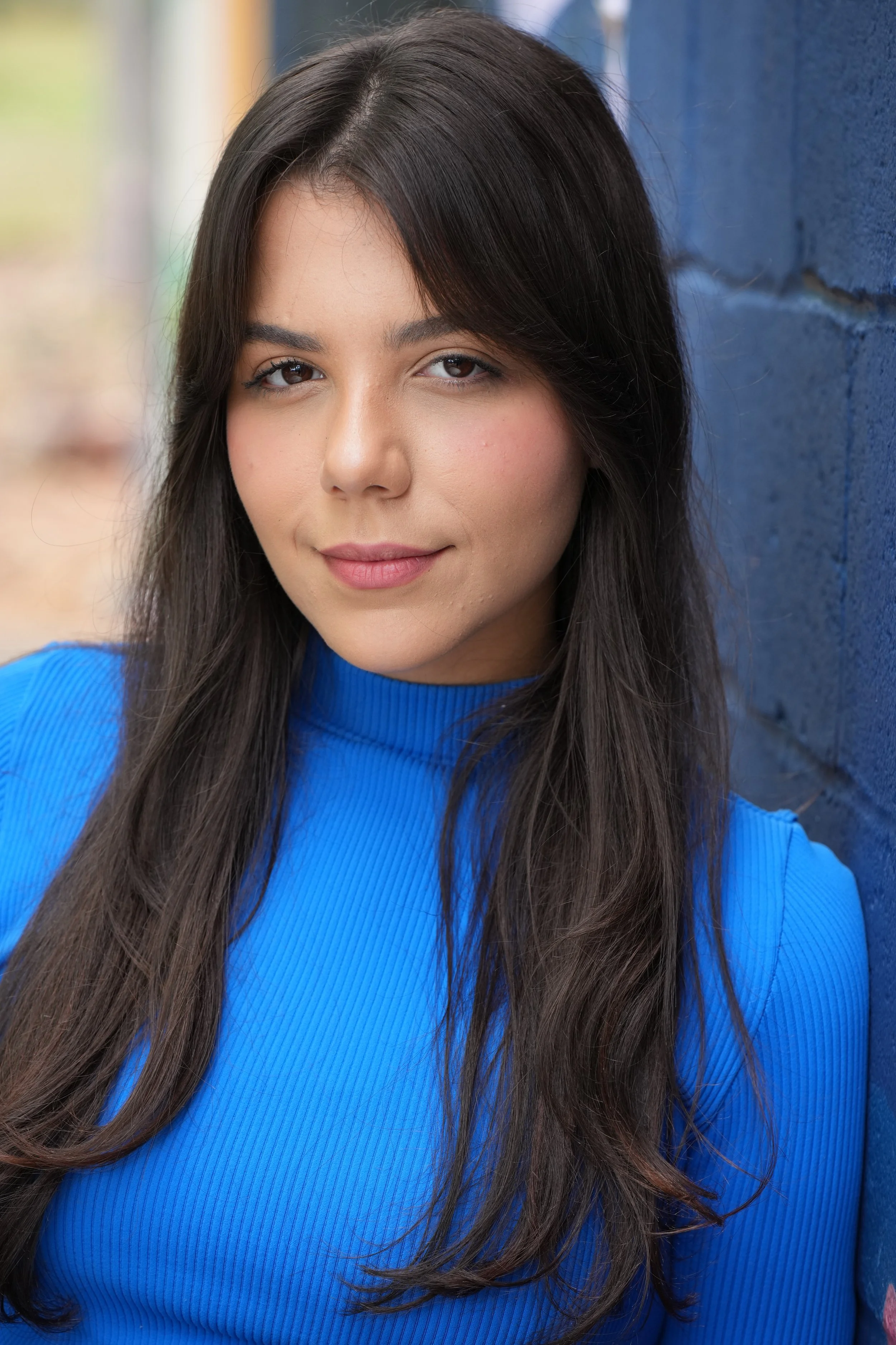 A young woman with long dark hair wearing a bright blue top, standing outdoors against a blue wall, with a blurred background.
