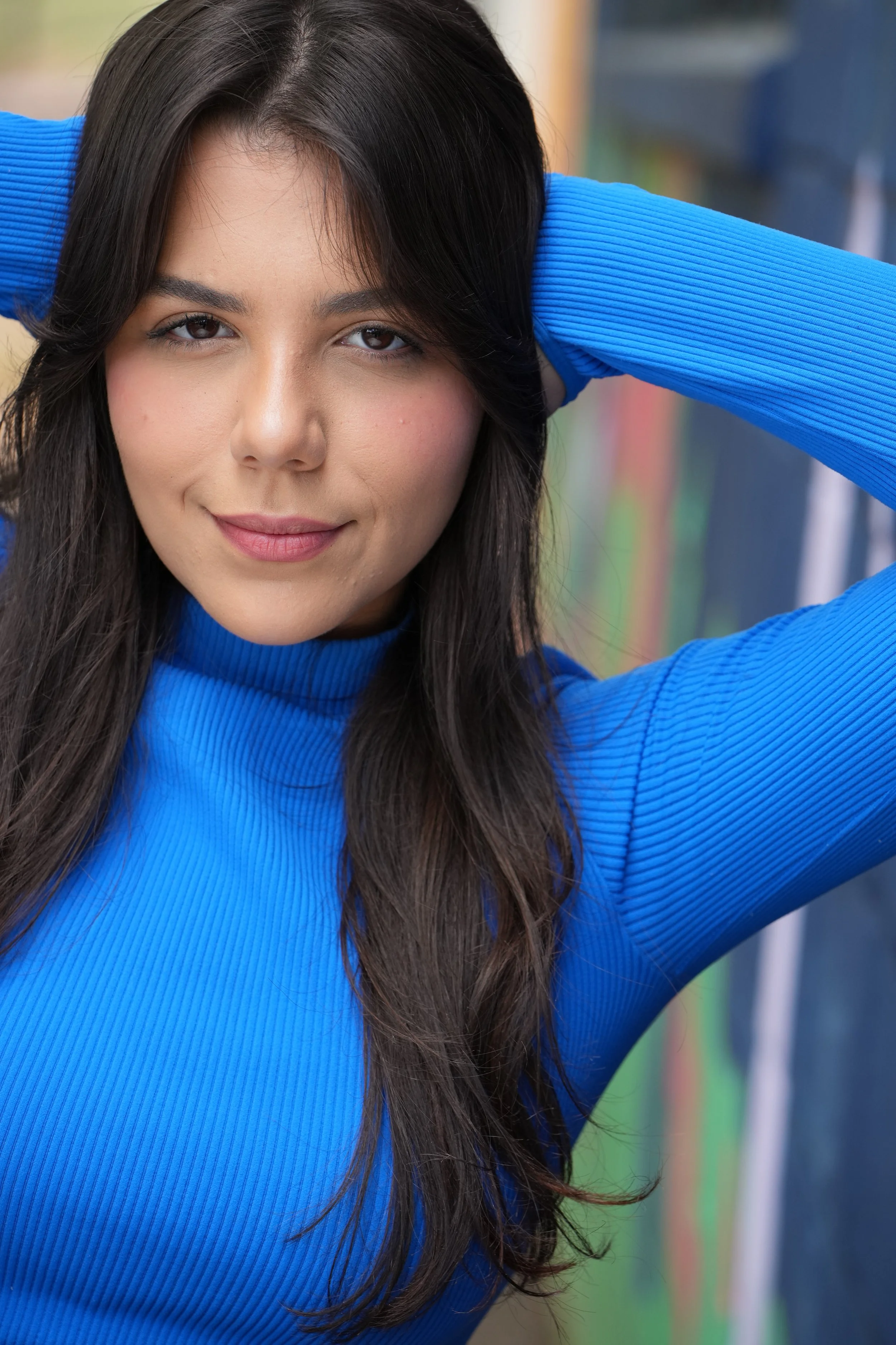 A woman with long dark hair, wearing a bright blue long-sleeve top, is smiling softly and posing with her hands behind her head. She has a natural makeup look and is outdoors, with a blurred colorful background.