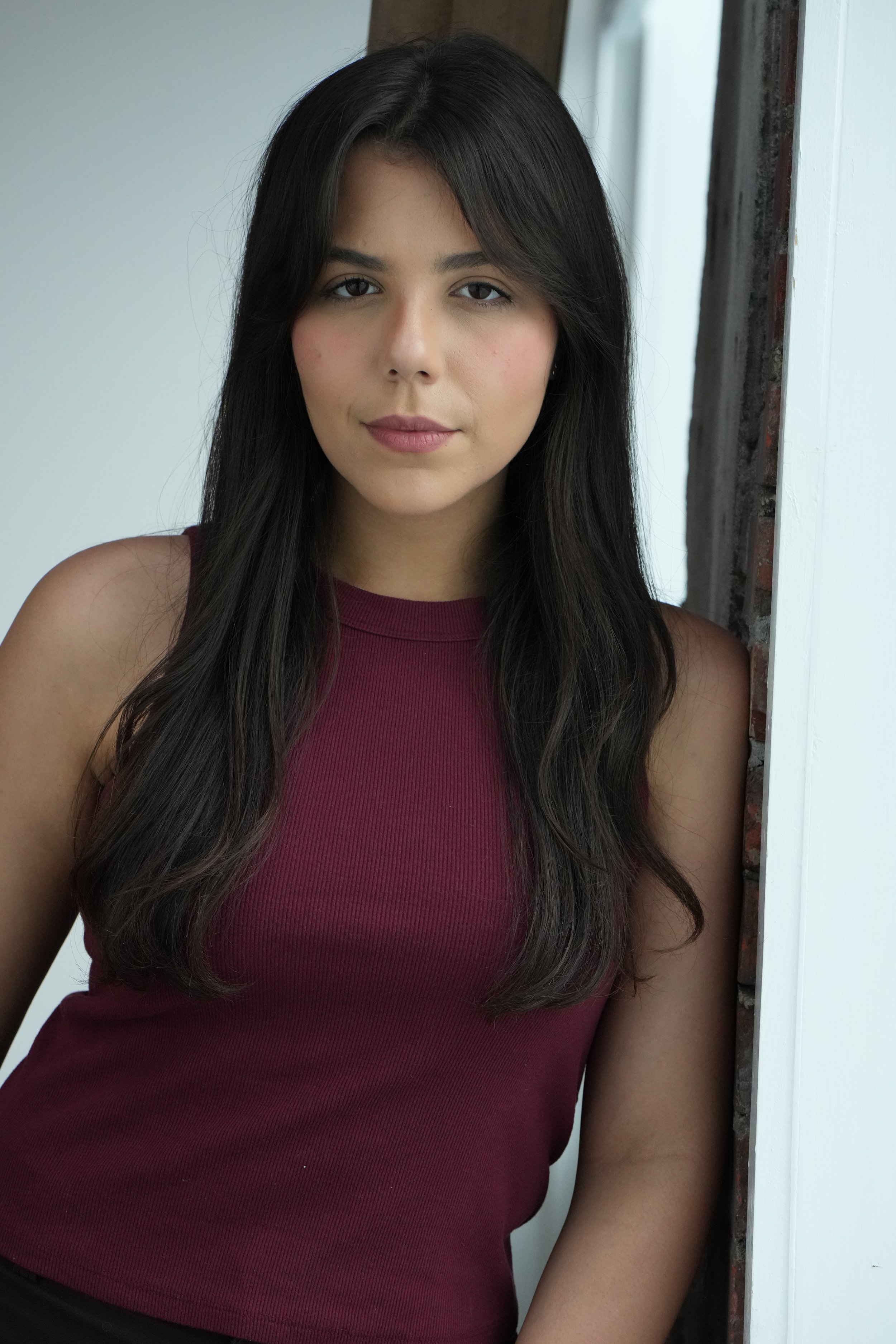 A young woman with long dark hair, wearing a sleeveless maroon top, standing indoors near a window with white trim and exposed brick wall.