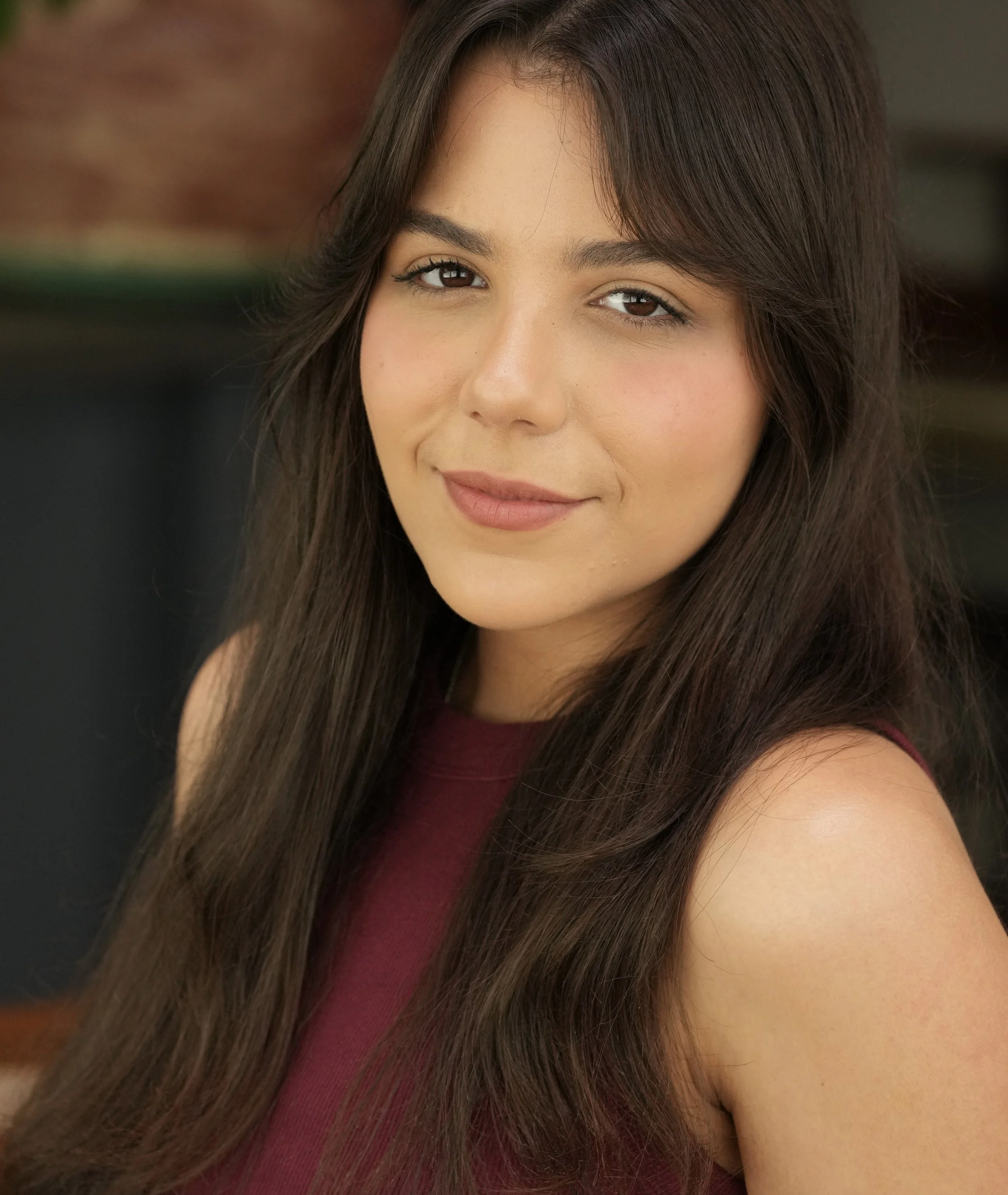 Close-up of a woman with long brown hair wearing a sleeveless maroon top, smiling softly with her face slightly turned to the side.