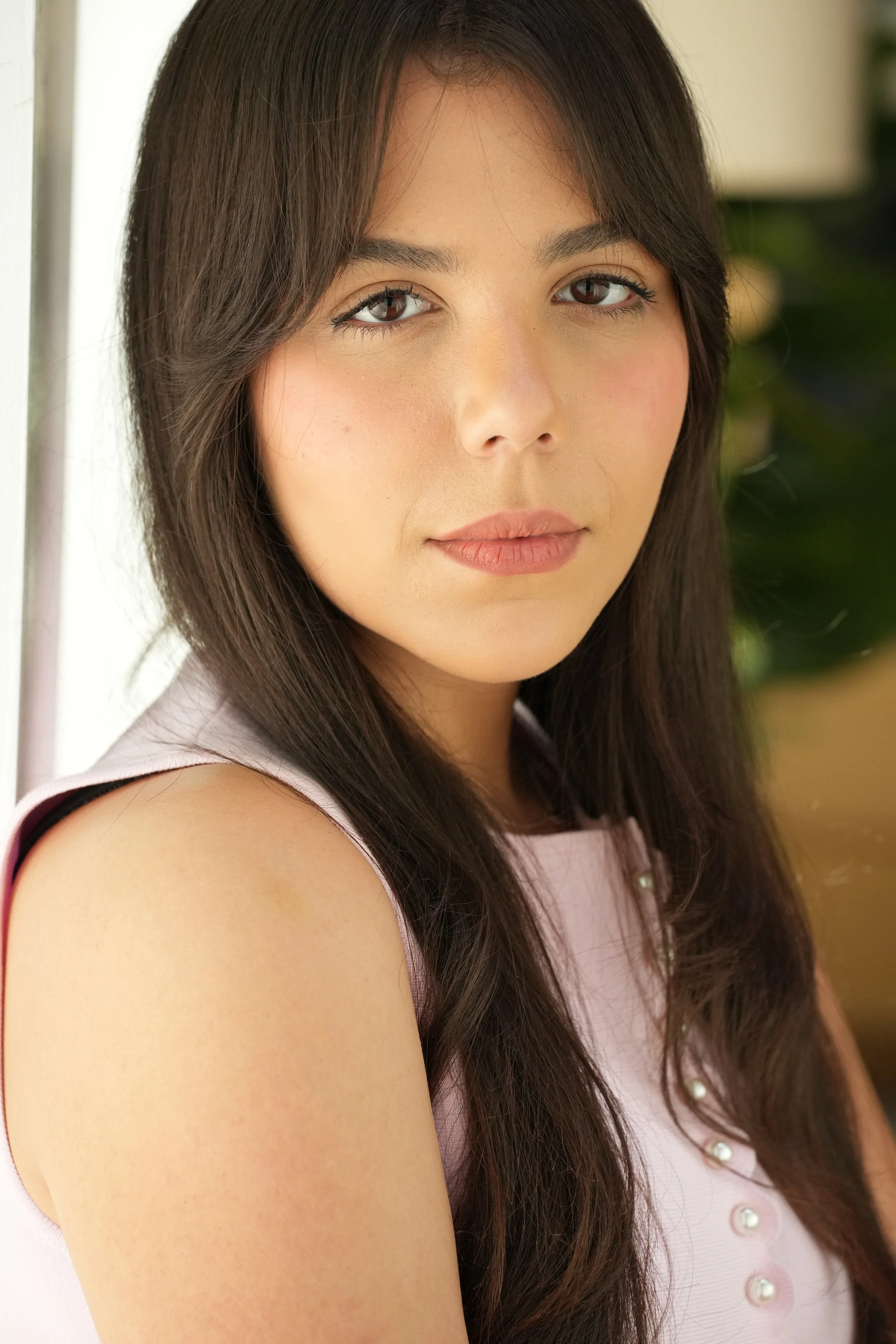Close-up of a young woman with long dark hair, wearing a light pink sleeveless top with pearl buttons, looking at the camera with a neutral expression.