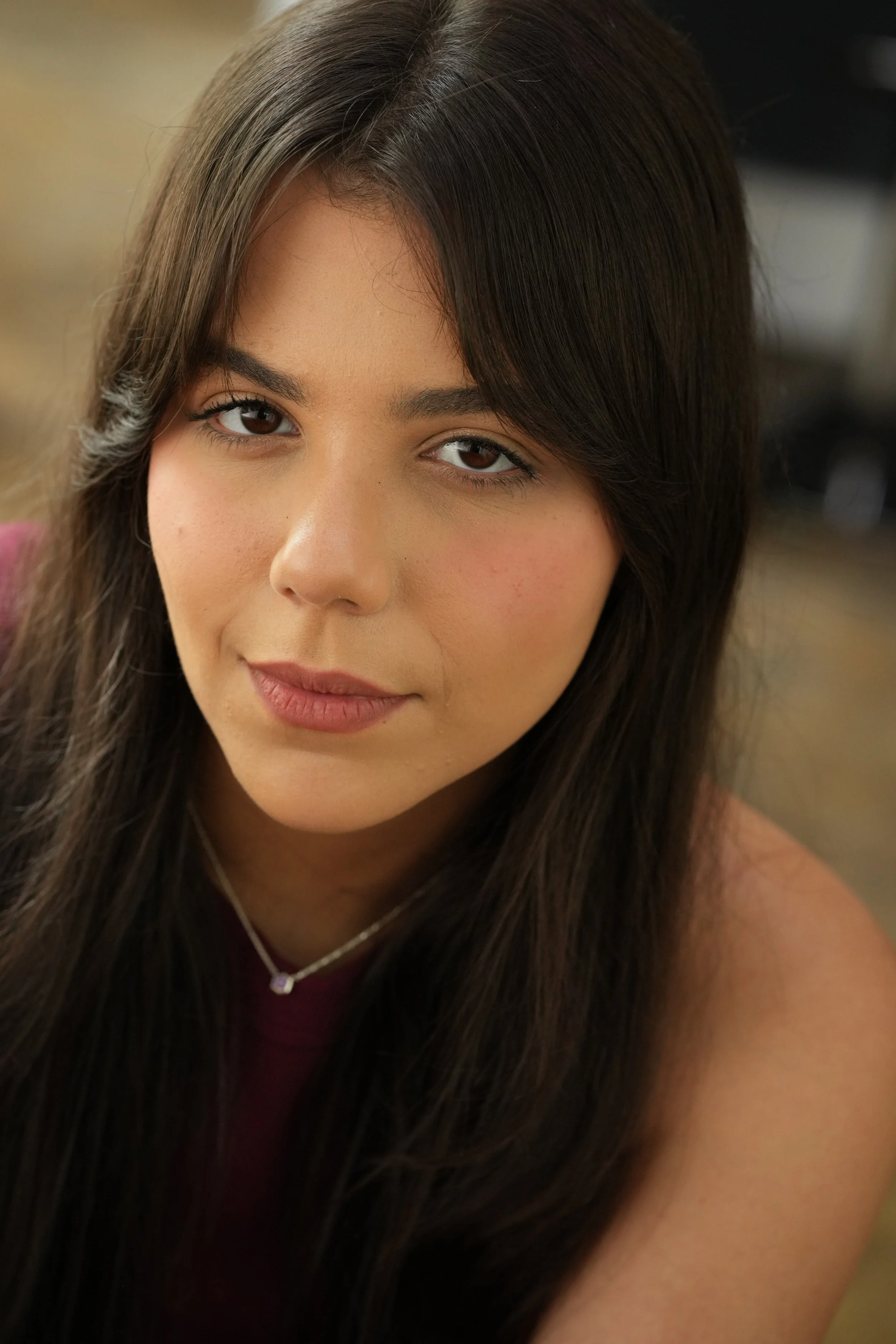 Close-up of a woman with long, dark hair, brown eyes, and a subtle smile, wearing a red top and a silver necklace, indoors.