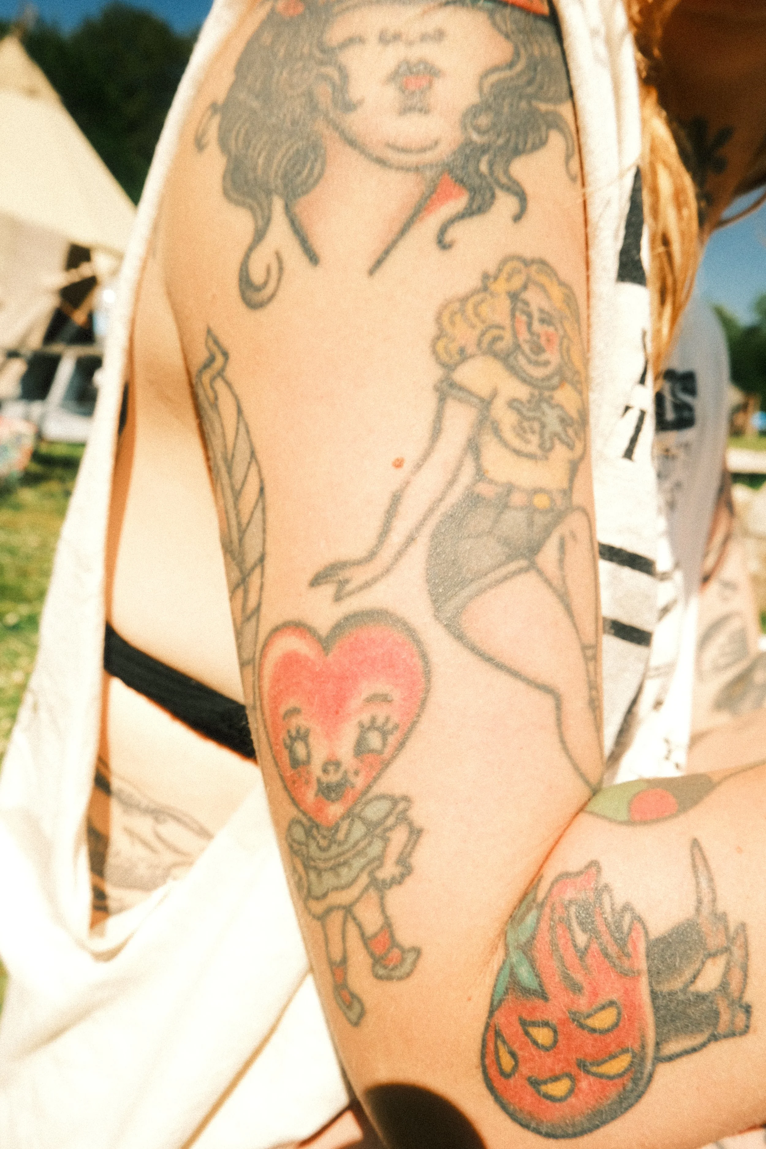 Tattoo of a woman with curly hair and a confident expression, holding a popsicle stick. On the forearm, there is a tattoo of a heart with a face, big eyes, and a wide smile. The person is wearing a short-sleeved shirt.