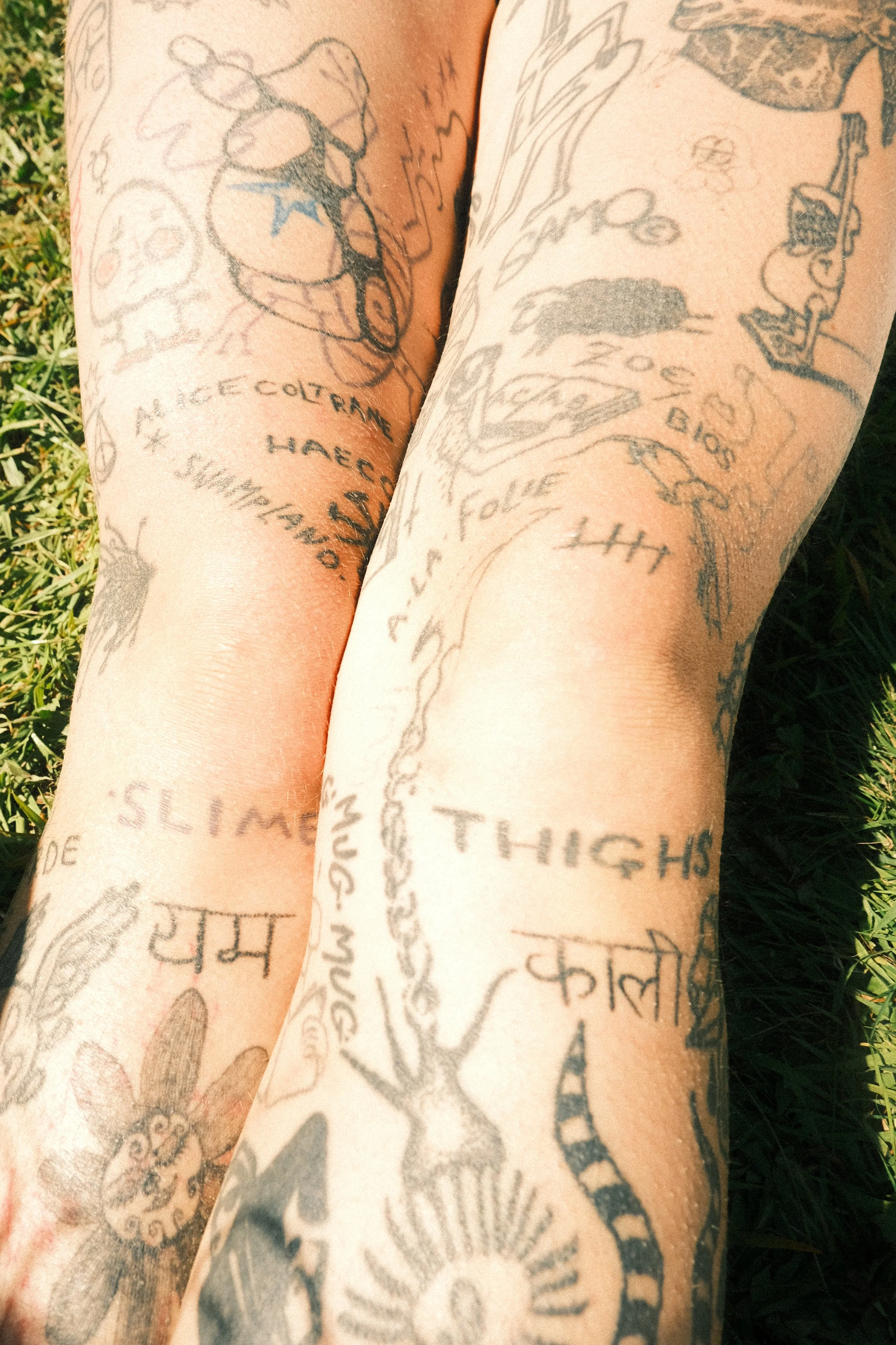 Close-up of a person's legs covered in various tattoos, with grass in the background.