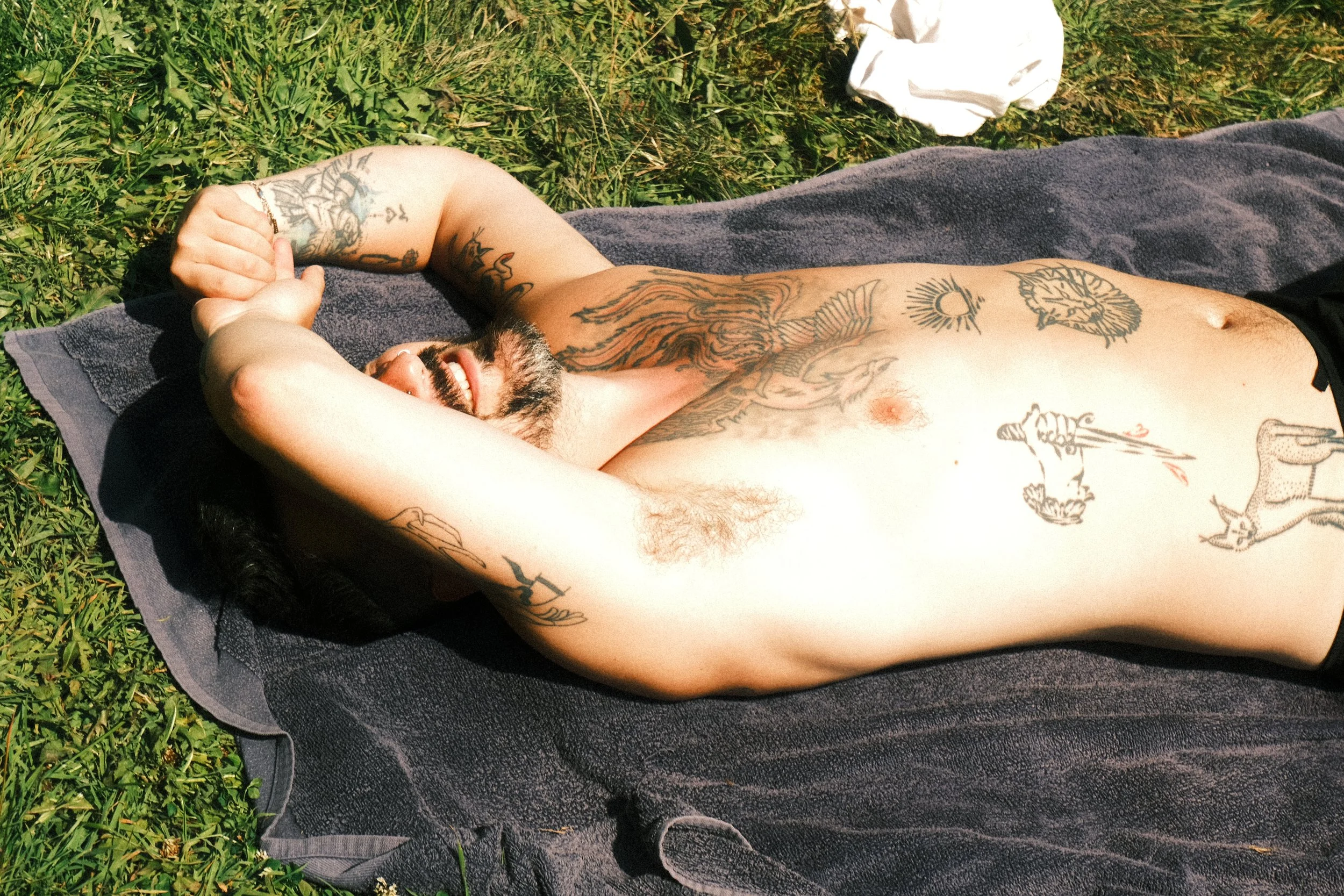A shirtless man with tattoos lying on a black towel outdoors on grass, smiling and posing with his arms behind his head.