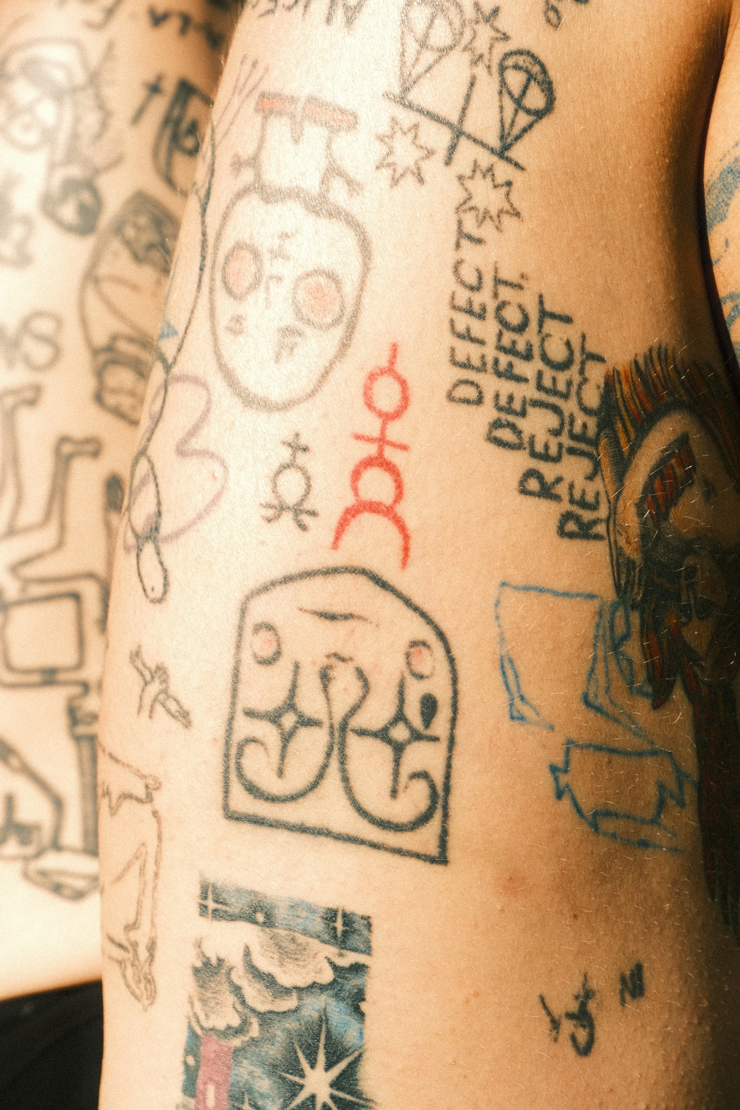 A close-up of a person's arm with multiple tattoos, including two skulls, a red barcode, and various symbols and text