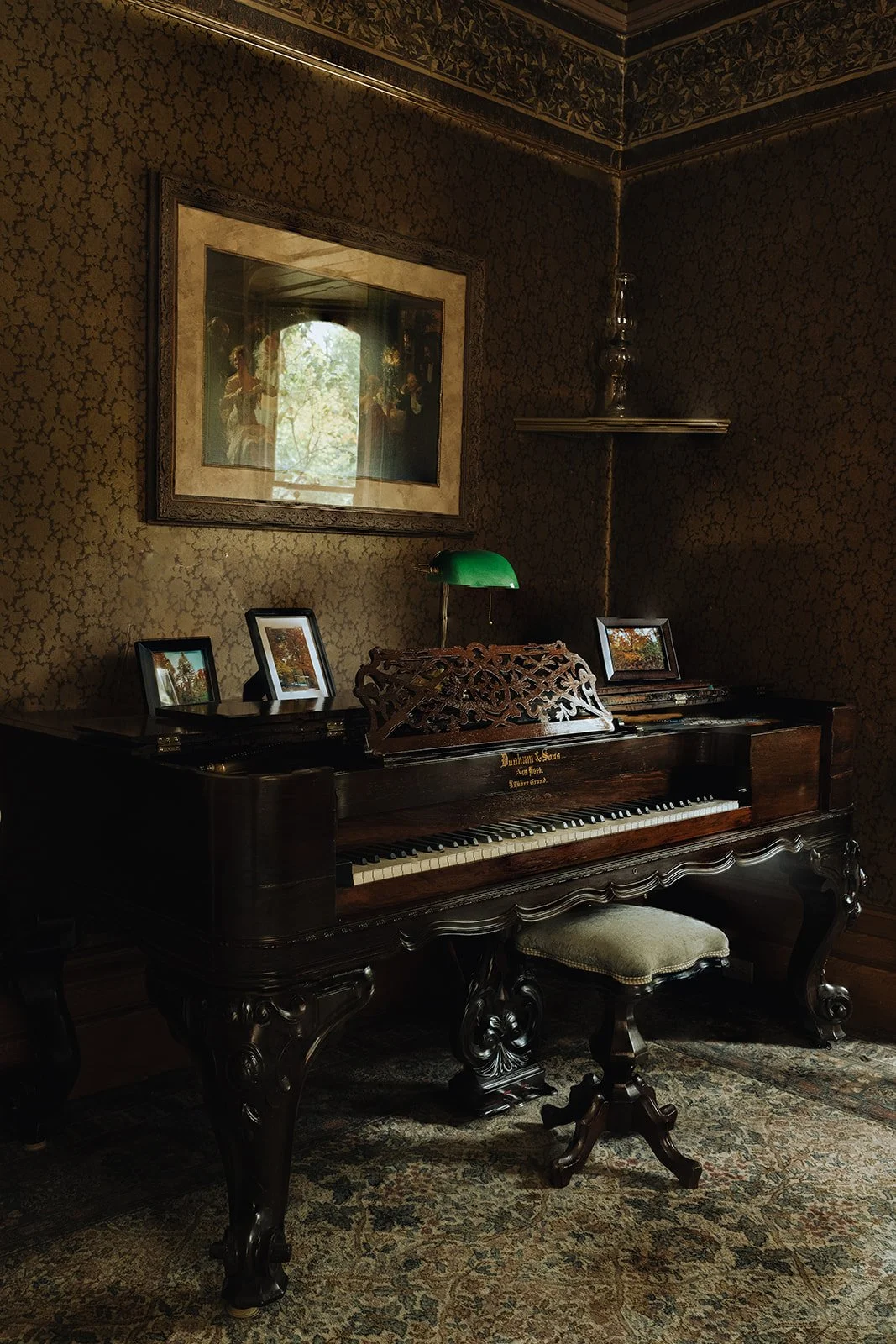 A vintage wooden upright piano with ornate legs and a carved music stand, situated in a dimly lit room with patterned wallpaper, four framed pictures on top, a green banker’s lamp, a stool with green upholstery, and a large framed artwork reflecting a window with trees outside.