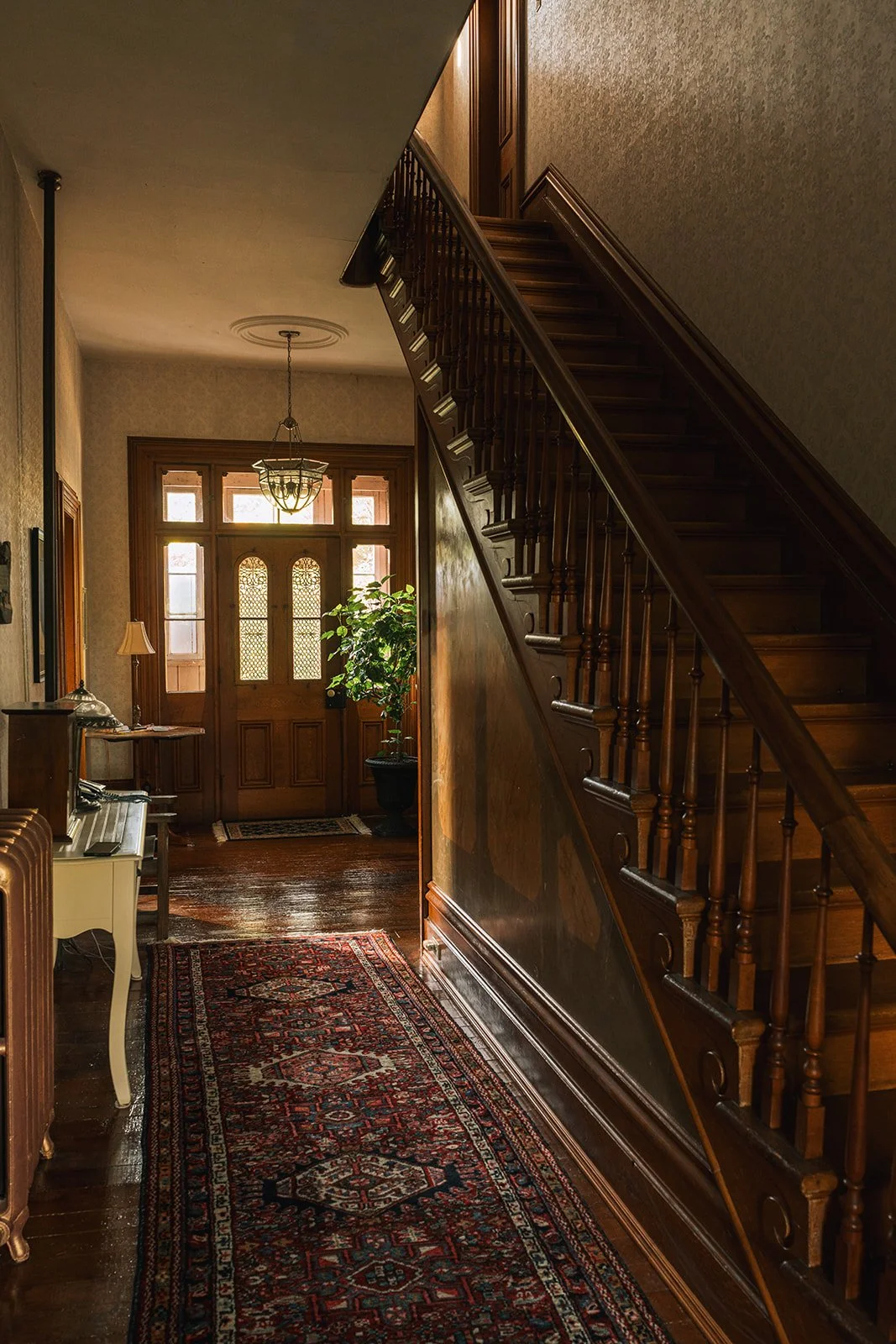 Entryway with wooden stairs, front door with stained glass, potted plant, patterned rug, and vintage furniture and decor.