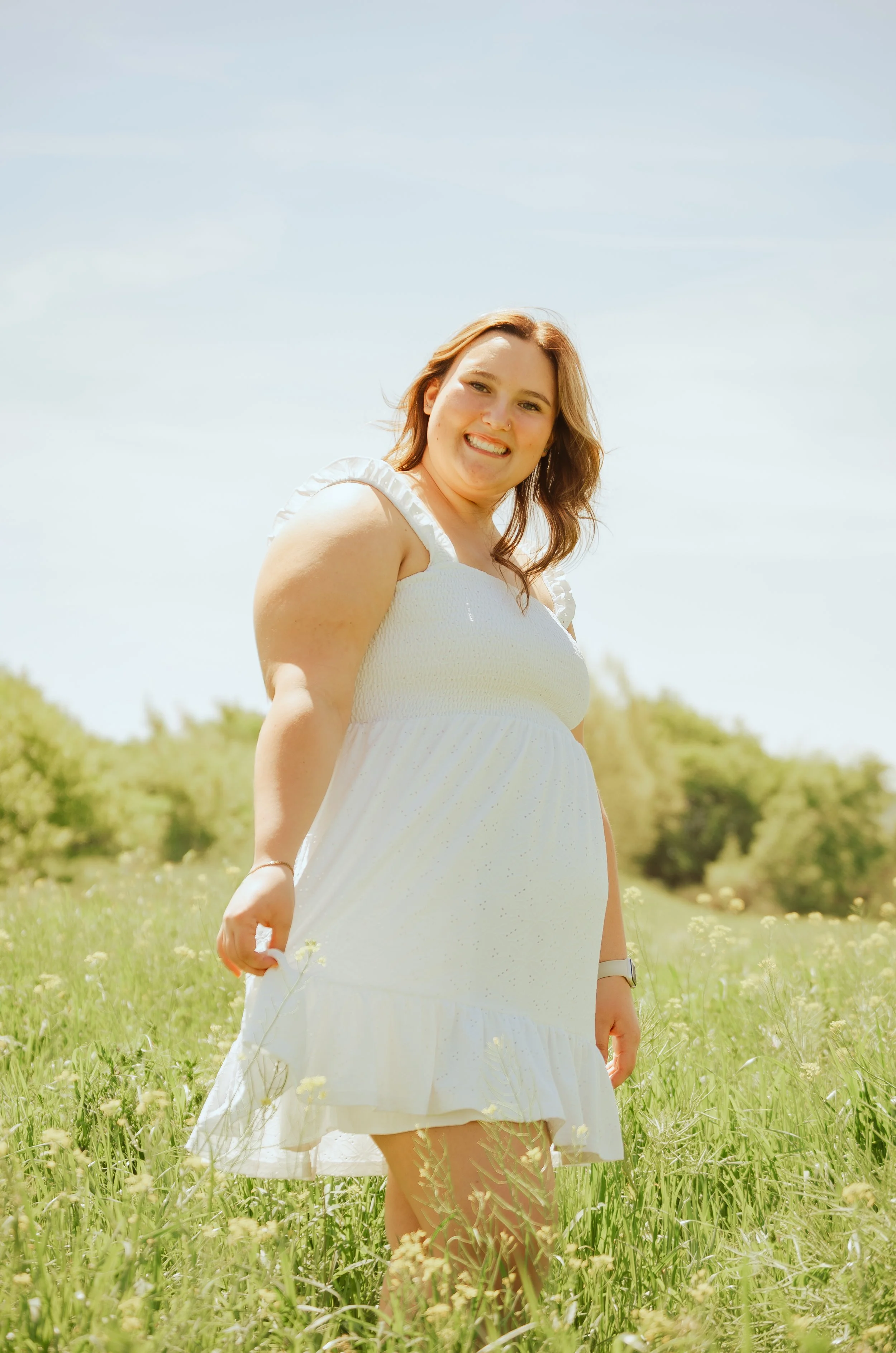 A woman with reddish-brown hair smiling, standing in a grassy field with small yellow and white flowers under a clear blue sky.