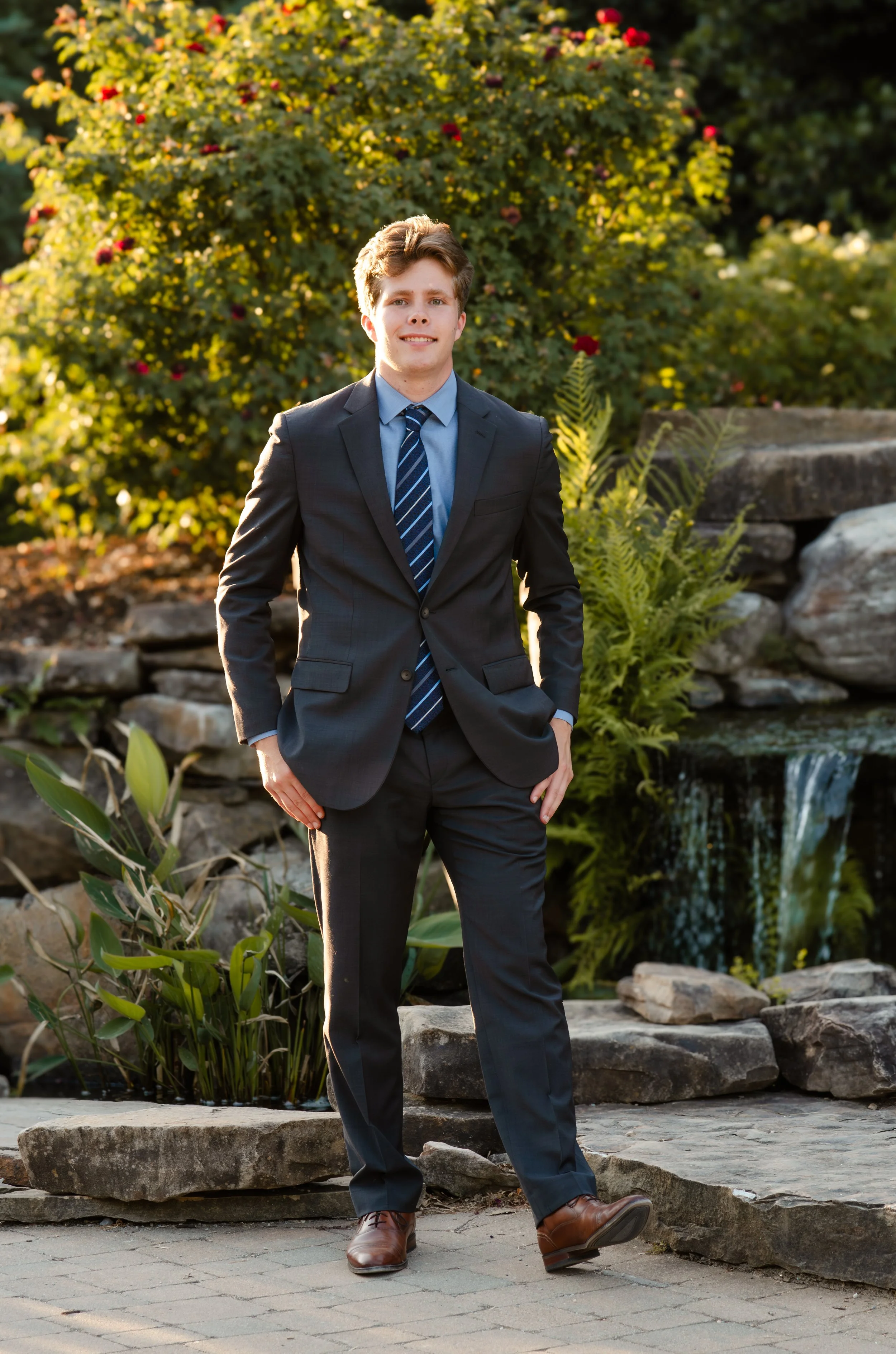 A young man in a dark suit, blue dress shirt, and striped tie standing outdoors on a stone path in front of a waterfall and lush greenery, smiling at the camera.