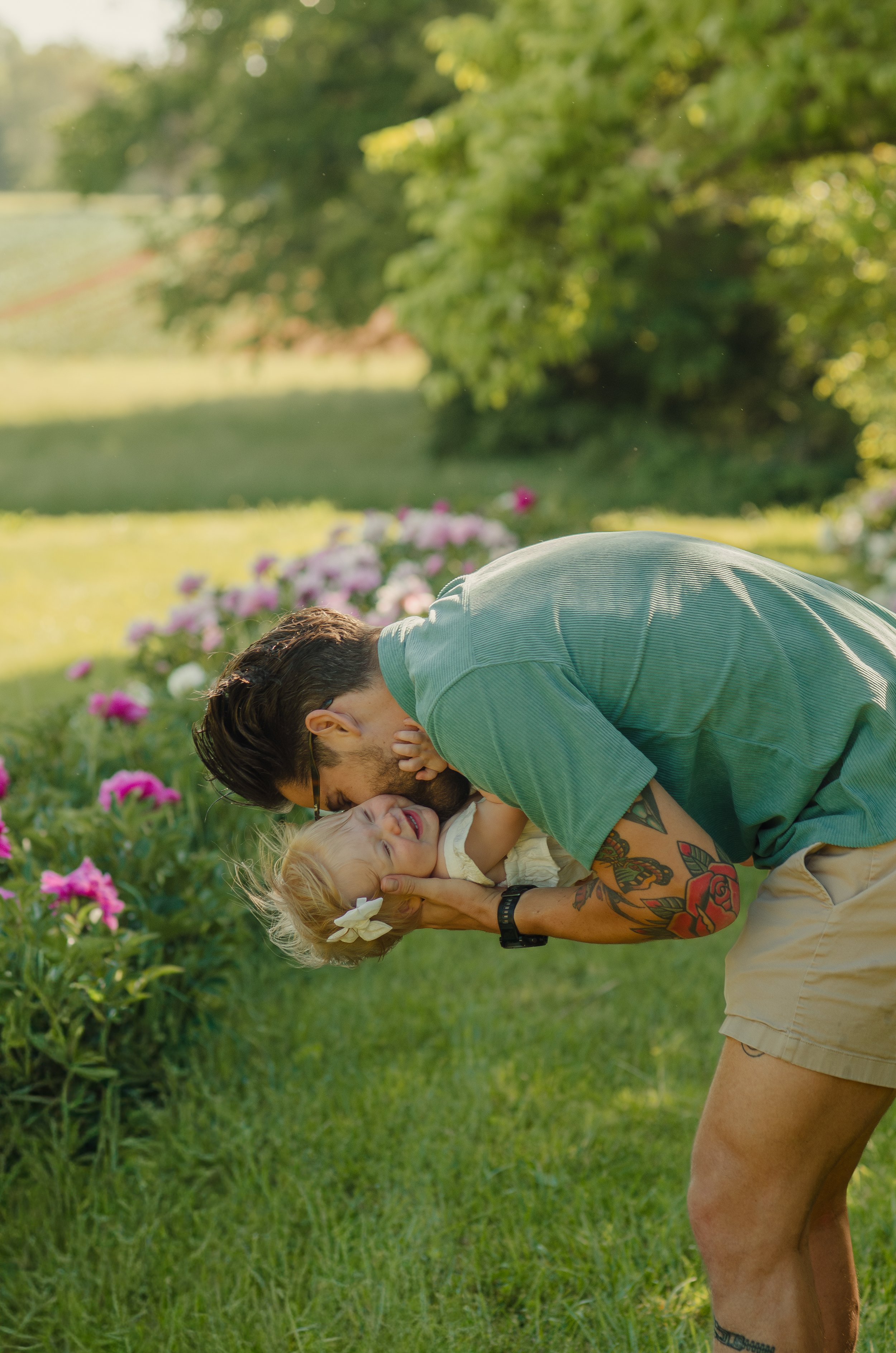 A man with tattoos on his arm and a young girl with blonde hair and a floral hair accessory are playing and kissing outdoors in a garden with pink flowers and green trees in the background.